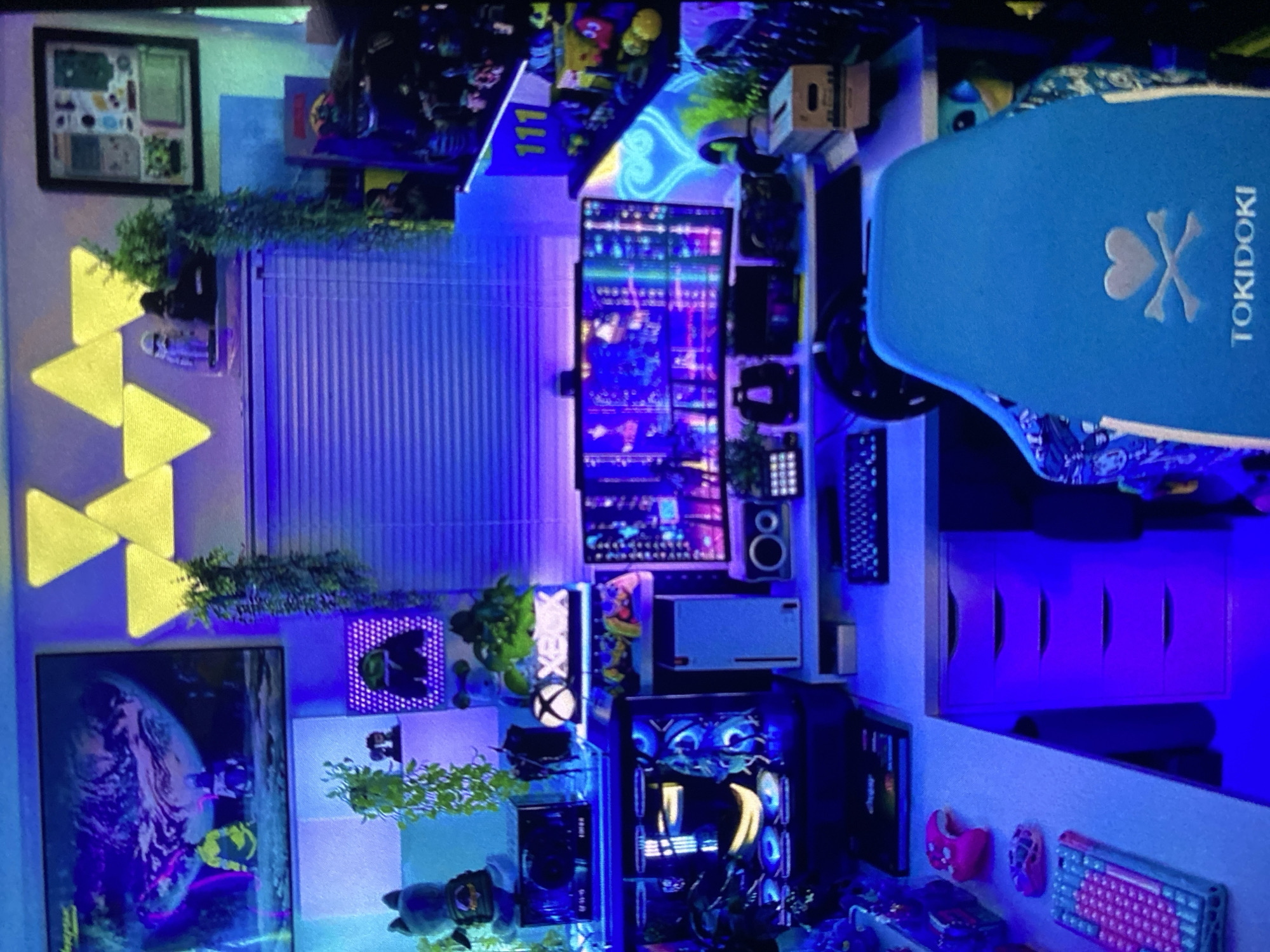 The image shows a highly decorated room, with a strong emphasis on gaming and computer equipment. Multiple monitors are stacked vertically, suggesting a multi-display setup. There's a gaming PC with illuminated fans and components, and a keyboard. The room is lit with blue and yellow ambient lighting, including triangular light panels on the wall. There are various decorations, including framed art, plants, and figurines. There is a skateboard with the word "TOKIDOKI" written on it. There is no information about location in Lhokseumawe, Indonesia, or any people.