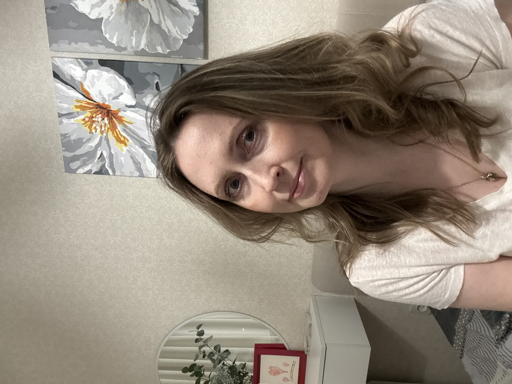 A woman with long, wavy light brown hair, fair skin, and light-colored eyes is captured in an indoor selfie, looking directly at the camera with a soft, gentle smile. She is wearing a simple, light-colored (likely white or off-white) t-shirt and a delicate gold necklace with a small pendant. Her body is positioned diagonally in the frame, suggesting a relaxed pose.
The background features two large, framed art pieces on a light, textured wall. These paintings depict stylized white flowers with bright orange and yellow centers against a grey background, resembling paint-by-number artwork. Below and to the right of the woman, a white piece of furniture is visible. On this surface, a round mirror reflects white horizontal blinds and a plant arrangement containing green leaves, possibly eucalyptus, and small white flowers. Next to the plant, there is a small red-framed picture or card with an abstract heart-like drawing.
The setting appears to be a domestic interior in Chaikovsky, Russia, possibly a living room or bedroom, characterized by soft, even lighting. No discernible text is visible in the image.