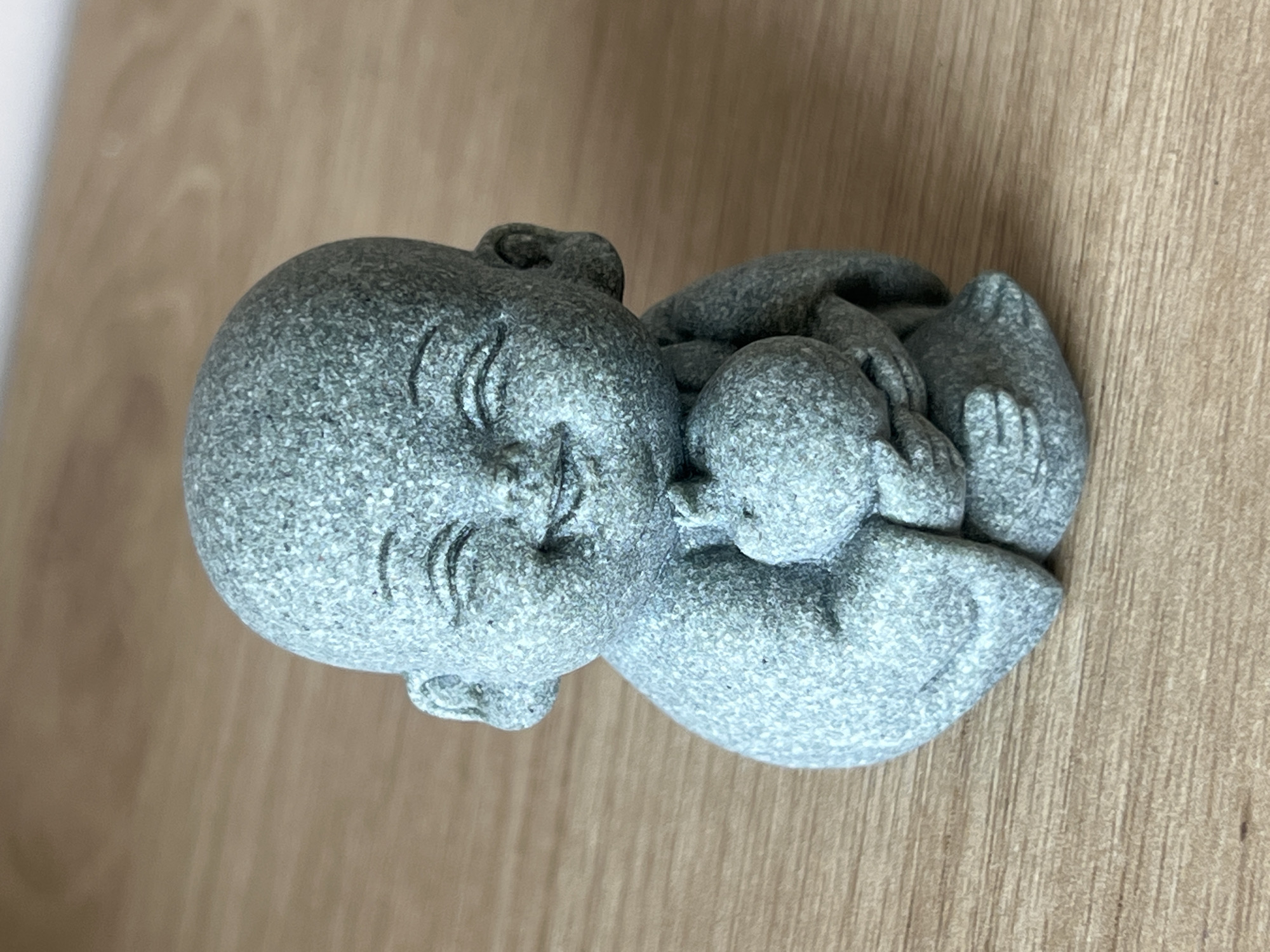 A gray stone statue of a smiling, reclining Buddha-like figure holding a smaller figure, is laying on a wood surface. The environment is neutral, offering no specific clues about the location or the Nam Natural Heritage Area Trang, Vietnam.