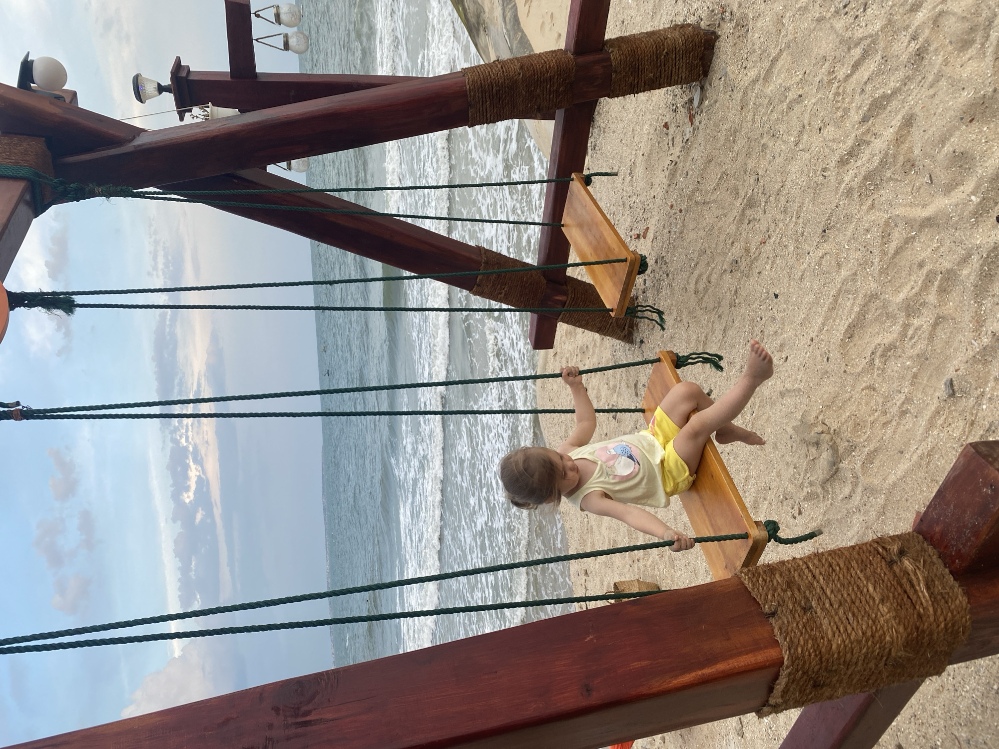 A young child in a yellow outfit is on a swing at the beach in Phu Thuy Ward, Vietnam. The swing is made of wood and rope. The ocean is in the background, and the sky is cloudy. The swing structure is made of dark wood, reinforced with ropes, and sits on the sand near the water's edge.