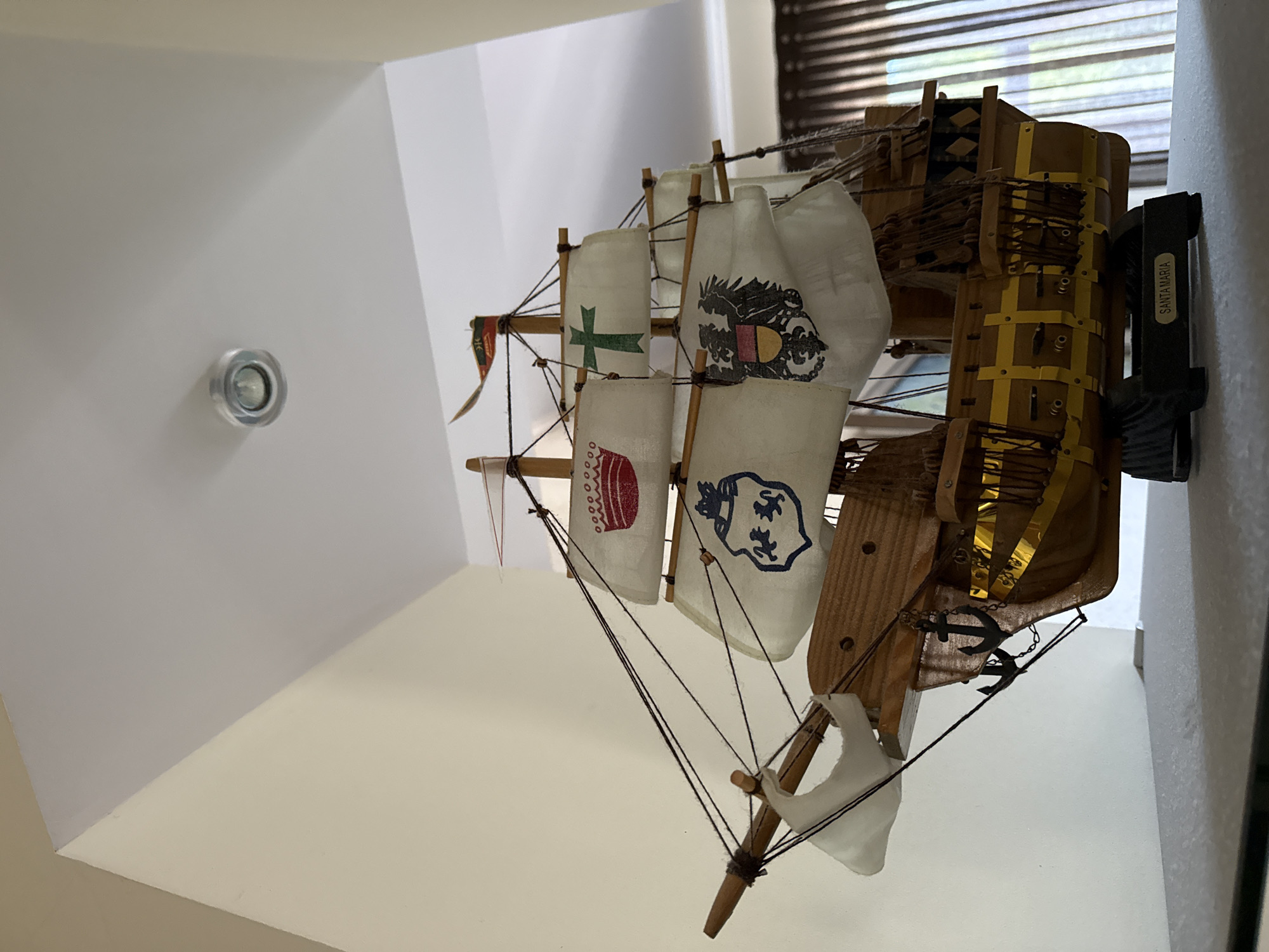 This media file depicts a model of the Santa Maria ship, likely a display piece. The model is predominantly made of wood, with white fabric sails that feature various emblems and designs. One sail has a green cross, another displays a coat of arms with an eagle, and a third shows a blue crest with lions. The ship has intricate rigging made of dark string, and two small anchor ornaments are attached to its side. The hull of the ship is detailed with yellow banding. The model is shown mounted on a white wall, with a black bracket labeled "SANTA MARIA" visible on the right side. The background includes a window with blinds and what appears to be a light fixture on the white wall to the left. The scene is indoors and well-lit. The overall impression is that of a well-crafted decorative item.