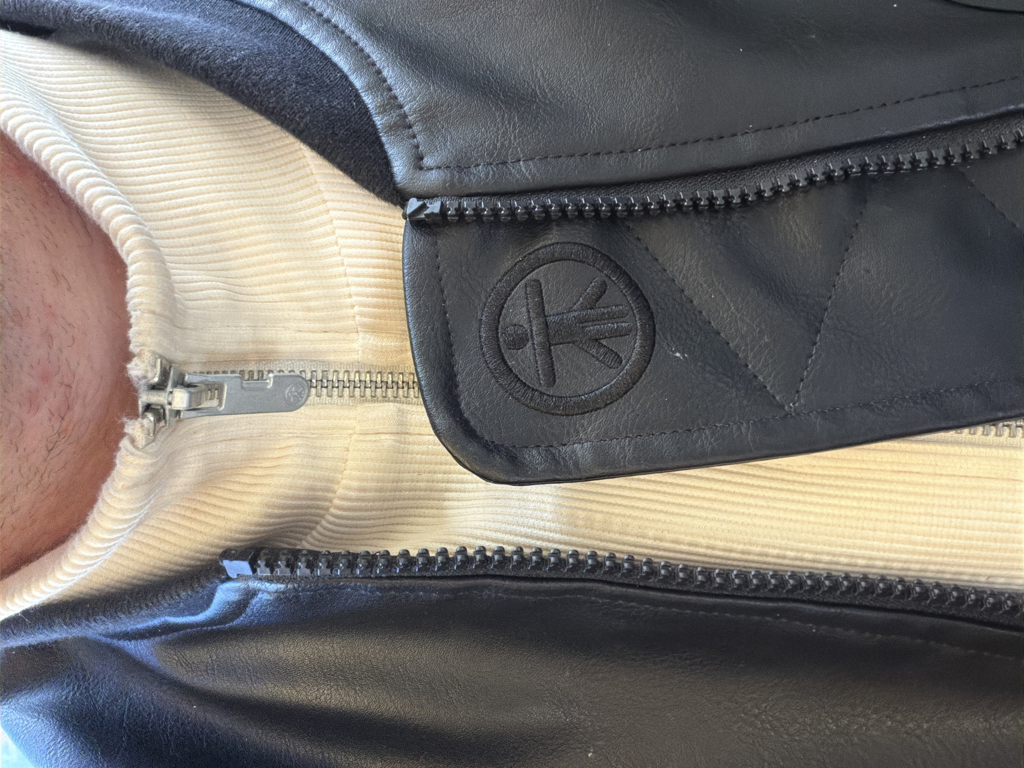 A close-up image depicts the upper torso of an individual wearing an off-white, ribbed turtleneck sweater partially unzipped at the neck, revealing a silver-toned metal zipper pull and teeth. Over the sweater, a black bomber-style jacket is worn, featuring a black zipper running down the center. On the viewer's left side of the jacket, a circular, black embroidered or embossed emblem depicts a stylized human figure with splayed limbs within a circle. Adjacent to this emblem, a section of the jacket exhibits diagonal or diamond-patterned stitching. No background or environmental details are visible beyond the subject's clothing. The stated location of this scene is London, United Kingdom; however, the image itself provides no visual information to confirm this.