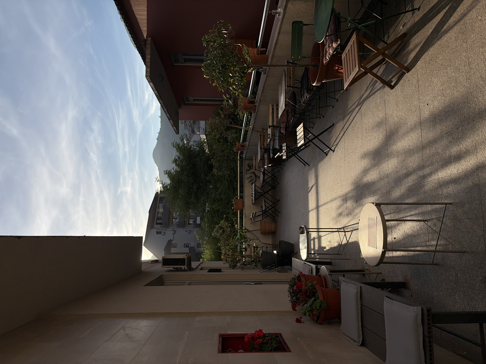 This image captures an outdoor terrace or balcony, likely part of a cafe or restaurant, rendered in the warm light of either early morning or late afternoon.
The scene is dominated by various seating arrangements. In the foreground on the right, there are built-in benches with light-colored cushions, adorned with terracotta pots containing vibrant red flowering plants, one of which has fallen over. A red recessed alcove in the adjacent wall also holds red flowers. Further along the terrace, numerous tables and chairs are neatly arranged. These include round and square tables, some with metal frames, accompanied by folding wooden chairs and metal chairs. One specific round table displays a sign that clearly reads "Cocktails."
To the left, a prominent red-painted building with windows is partially visible. Beyond the railing that borders the terrace, a panoramic view unfolds, featuring distant mountains under a wide, clear blue sky streaked with wispy white clouds. Green trees are interspersed among other residential buildings in the mid-distance. The sun, low in the sky, casts long, defined shadows across the textured floor of the terrace, creating a striped pattern and highlighting the contours of the furniture.
No people are visible, lending a peaceful and tranquil atmosphere to the inviting outdoor space. The weather appears fair and pleasant.