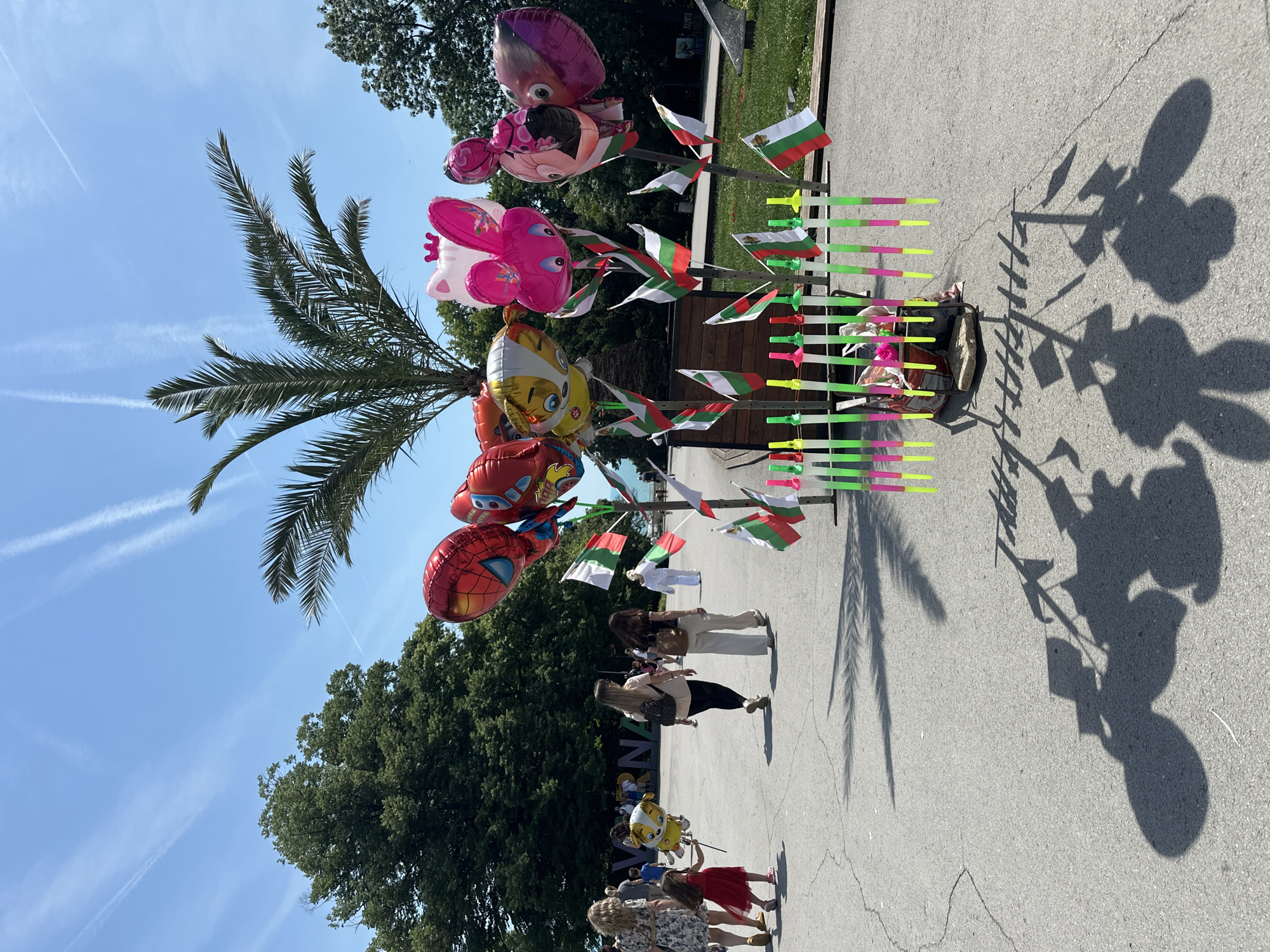 This image captures an outdoor scene on a sunny day in the Sea Garden of Varna, Varna Province, Bulgaria. The central feature is a vendor's improvised stand situated under a palm tree on a paved walkway.
The stand displays various items for sale:
* Multiple large, helium-filled foil balloons depicting popular characters, including a red Spider-Man, a yellow cartoon dog (possibly from Paw Patrol), a pink cat with a crown (resembling Hello Kitty), and a pink bunny.
* Numerous small Bulgarian flags (horizontal white, green, and red stripes) are attached to sticks and arranged around the balloons.
* Approximately twelve fluorescent plastic light-up toy swords or glow sticks, colored green, pink, and yellow, are standing upright in a base at the bottom of the display. A dark bucket or container is visible within the base of this section.
The ground is paved asphalt, showing some cracks. Distinct shadows of the palm tree, balloons, and flag sticks are cast onto the pavement, indicating direct sunlight from an elevated position.
Several people are walking on the paved path. In the foreground, a woman in a floral dress holds the hand of a young child in a red dress; the child carries a yellow character balloon on a stick. Further back, two women are walking away from the camera. One wears a light-colored top and dark pants with a backpack, while the other wears a dark top and light-colored wide-leg pants with a shoulder bag. In the distance to the right, a person in a white, full-body suit is also walking away.
In the mid-ground, large, white, freestanding letters spell "VARNA," confirming the city. Beyond this sign, a strip of light blue water, likely the Black Sea, is visible. The background features lush green deciduous trees to the left and right, and a bright blue sky with multiple prominent white contrails. A dark, angular sculpture is partially visible on the paved area to the far right.