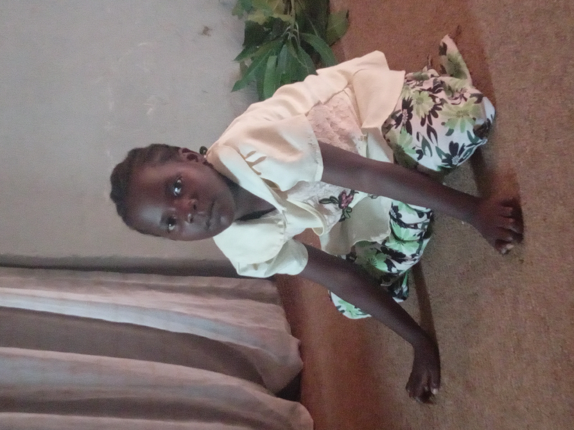 A young, dark-skinned girl with braided hair is kneeling on a light brown floor. She wears a light yellow blouse with a lace inset, and patterned pants with green and white floral designs. Her arms are extended forward, and she is looking directly at the camera. A piece of green foliage is visible in the upper background. There is a partially visible bed or fabric draped on a structure to the left.
The provided location (Sabon Tasha, Nigeria) is assumed true but cannot be verified. There is nothing in the image itself that specifically indicates its location.
