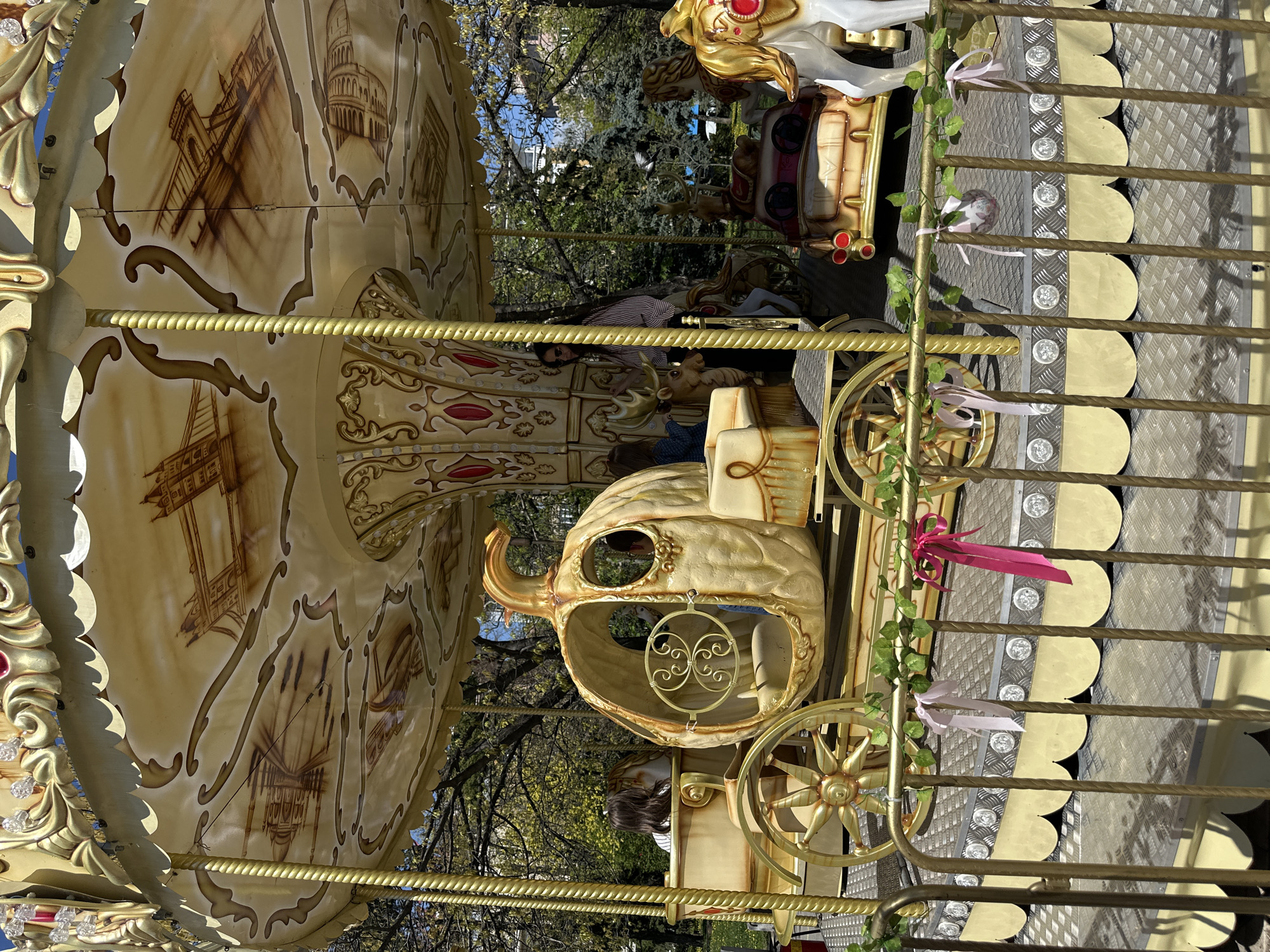 The image captures a portion of a carousel, featuring a decorative, cream-colored pumpkin carriage and ornate gold detailing. The carousel canopy displays sepia-toned images of architectural landmarks. The foreground shows gold metal bars, adorned with ribbons and artificial ivy. Through the carousel's structure, a blurred background reveals trees, suggesting an outdoor setting, likely a park or recreational area in Sofia, Bulgaria. There are people visible in the background.