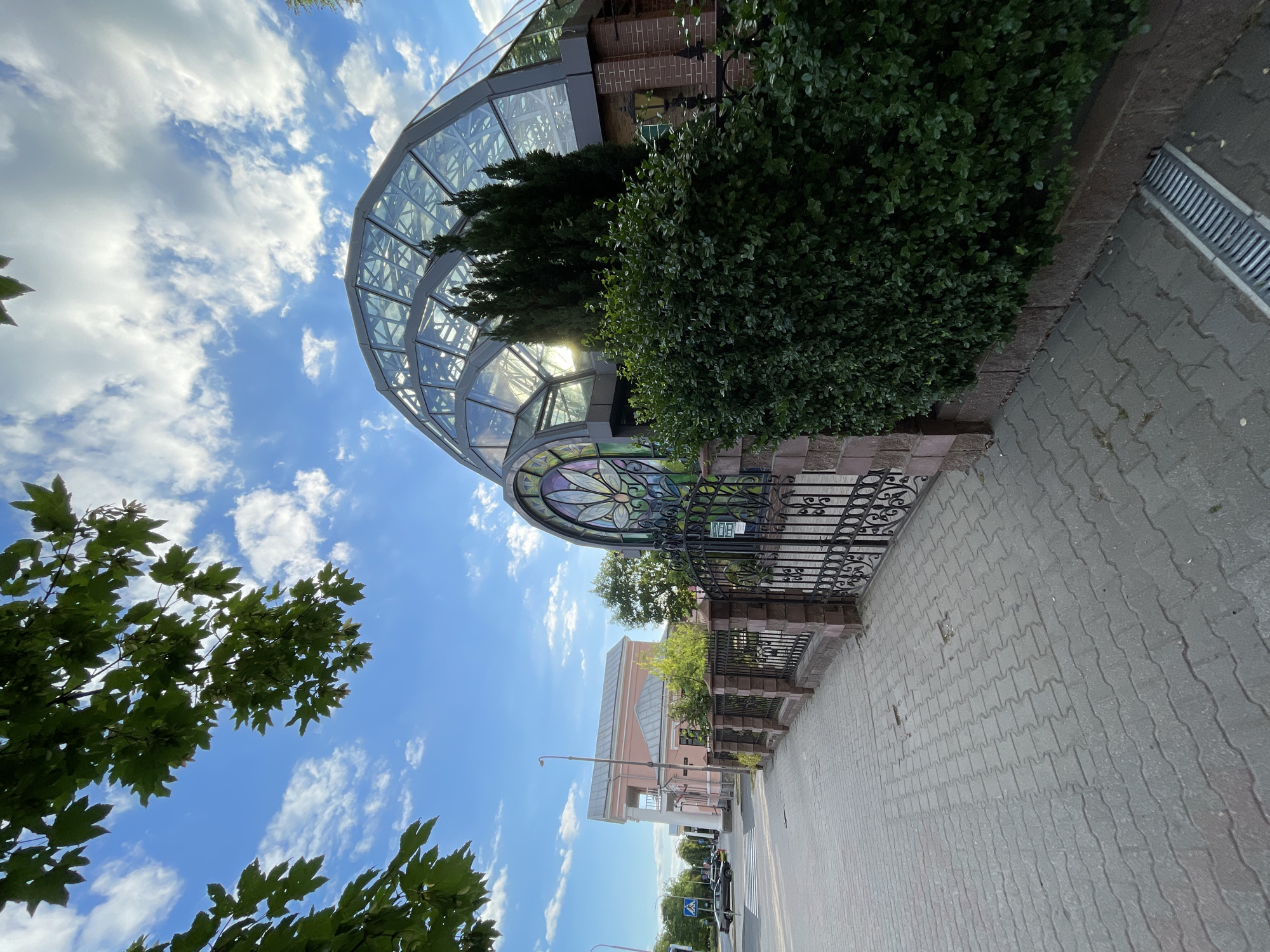 An urban street scene in Brest, Belarus, captured on a sunny day under a blue sky with scattered white cumulus clouds. In the foreground on the left, a grey interlocking paver sidewalk extends diagonally. A black wrought-iron fence with stone pillars runs parallel to the sidewalk on the right. A dense green hedge is situated behind this fence. A metal storm drain grate is visible on the sidewalk in the lower right corner.
To the right, behind the hedge, is a distinctive modern building featuring a large, multi-faceted glass dome roof structure that reflects the sunlight. An ornate archway on the left side of this building incorporates a colorful stained-glass window with a floral, lotus-like design. Adjacent to the archway, a section of the building's facade is constructed of reddish-brown brick, from which an antique-style black metal street lamp protrudes. Several tall, slender evergreen trees are positioned between the hedge and the glass dome. A green sign with white Cyrillic text indicating "Рэжым працы" (Working hours) is affixed to the fence gate, along with a smaller green sign on the brick wall.
In the midground on the left, a paved street is visible, along with a dark-colored sedan situated near a pedestrian crossing. A grey lamppost stands beside the street. Further in the background on the left, a two-story classical-style building with light pink walls, white columns, and a grey hip roof is present. This structure is identifiable as the main building of the Brest Regional Museum of Local Lore.
Green foliage from tree branches with lobed leaves is visible in the upper left and central top portions of the image, silhouetted against the sky. No human figures are distinctly visible.
This scene depicts the Winter Garden annex of the Brest Regional Museum of Local Lore, located on Karl Marx Street in Brest, Belarus.
