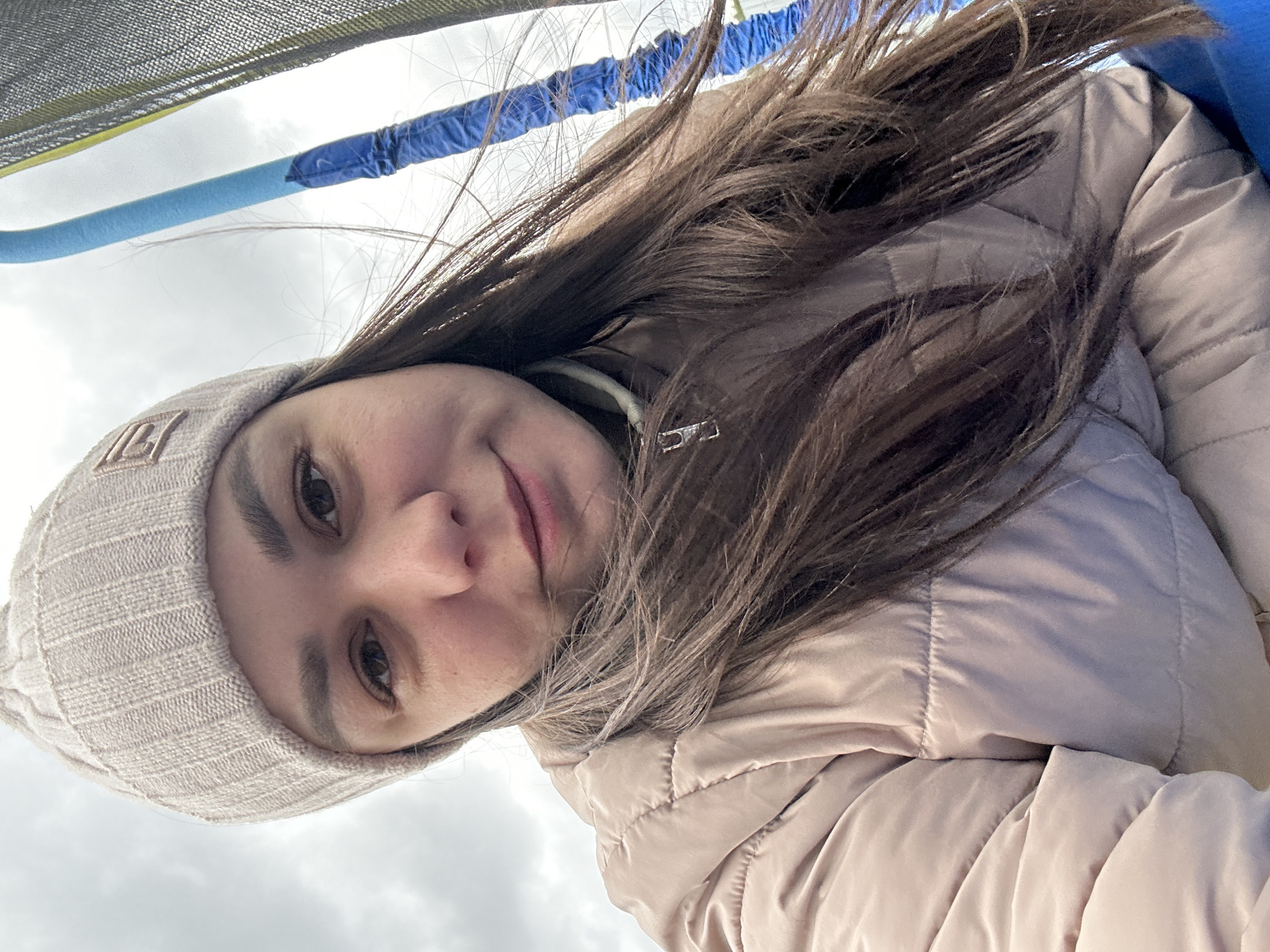 A young woman is captured in a close-up selfie, looking directly at the camera with a calm, slightly smiling expression. The image is rotated diagonally, with the woman positioned from the bottom-left to the upper-right of the frame.
She has long, dark brown hair, some strands of which appear windblown or disheveled. She is wearing a light beige or off-white knitted beanie hat with a distinct, possibly embroidered, logo on the front resembling two intertwined 'L's or a stylized monogram. Her outerwear consists of a light pinkish-beige puffer jacket, visible across her shoulders and arm.
The background is an overcast sky, suggesting a cloudy daytime setting. Above and behind her head, elements of a recreational structure are visible: blue and lighter blue padded bars or pipes, likely part of an enclosure, and a dark mesh net at the very top. These details indicate she is outdoors, possibly within or next to a trampoline or playground equipment in Chaikovsky, Russia.