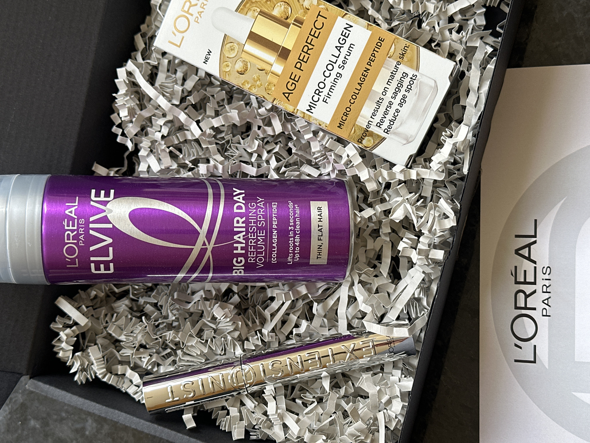 A top-down, close-up shot shows a black gift box filled with crumpled, silver-white shredded paper. Inside the box are several L'Oréal beauty products. A purple and silver can of L'Oréal Elvive "Big Hair Day" refreshing volume spray sits alongside a gold-toned L'Oréal "Extensionist" mascara tube. Adjacent to these items is a white box of L'Oréal "Age Perfect" micro-collagen firming serum. A white information card with the L'Oréal Paris logo is also within the box. There are no people visible in the image. Based on the inclusion of L'Oréal Paris products sold by Boots, it is probable that this image was taken in the city of Tunbridge Wells, United Kingdom.