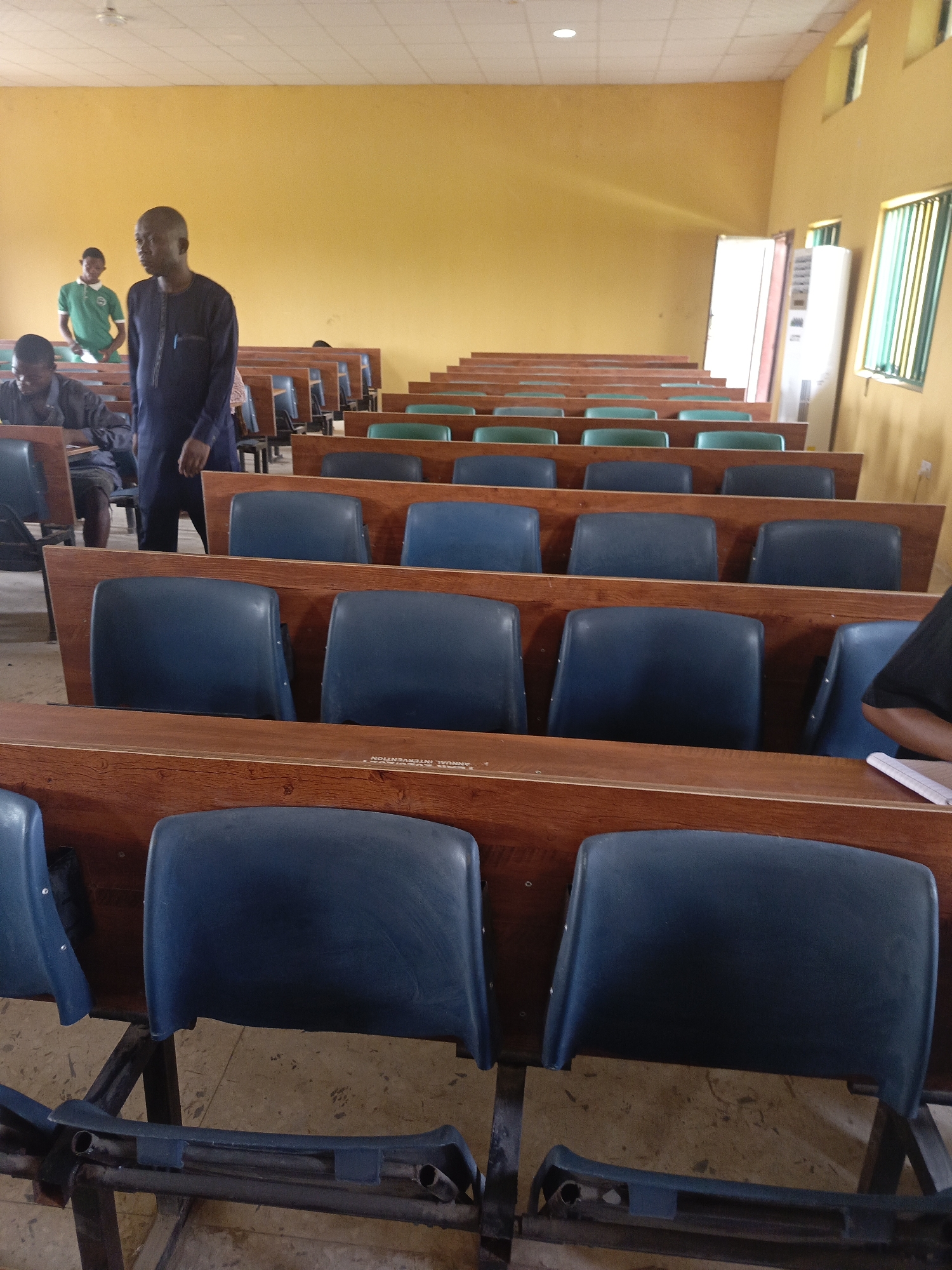 This image depicts the interior of what appears to be a lecture hall, classroom, or meeting room in Akwanga, Nigeria. The room is characterized by rows of tiered seating and attached wooden-textured desks. The majority of the chair backrests are dark blue plastic, though some in the upper rows appear to be a lighter green or teal. Several seats show signs of wear, with some of the seat bottoms missing or damaged.
In the foreground, a man wearing a dark blue traditional long-sleeved tunic (possibly a kaftan or similar garment) stands in an aisle, looking towards the right side of the room. A pen is visible in his chest pocket. To his left, partially obscured by the desk, another man is seated, looking down, possibly writing or engaged with an item on his desk. Further back, on the left side of the room, a third man in a bright green polo shirt stands near a desk. On the far right, only a person's arm and shoulder are visible, along with a white notebook or binder on a desk, suggesting they are seated and possibly taking notes. Most of the seats in the room are unoccupied.
The walls of the room are painted a light yellow or ochre color. On the right side, several windows with green horizontal security bars allow natural light into the space, indicating it is daytime. A tall, white appliance, possibly an air conditioner or refrigerator, stands next to a doorway on the right. The ceiling consists of white acoustic tiles with a light fixture visible.
Visible text on one of the wooden desk surfaces reads "ANNUAL INTERVENTION". The overall atmosphere is quiet and functional, with the sparse occupancy suggesting either preparation for an event, a break, or a sparsely attended session.