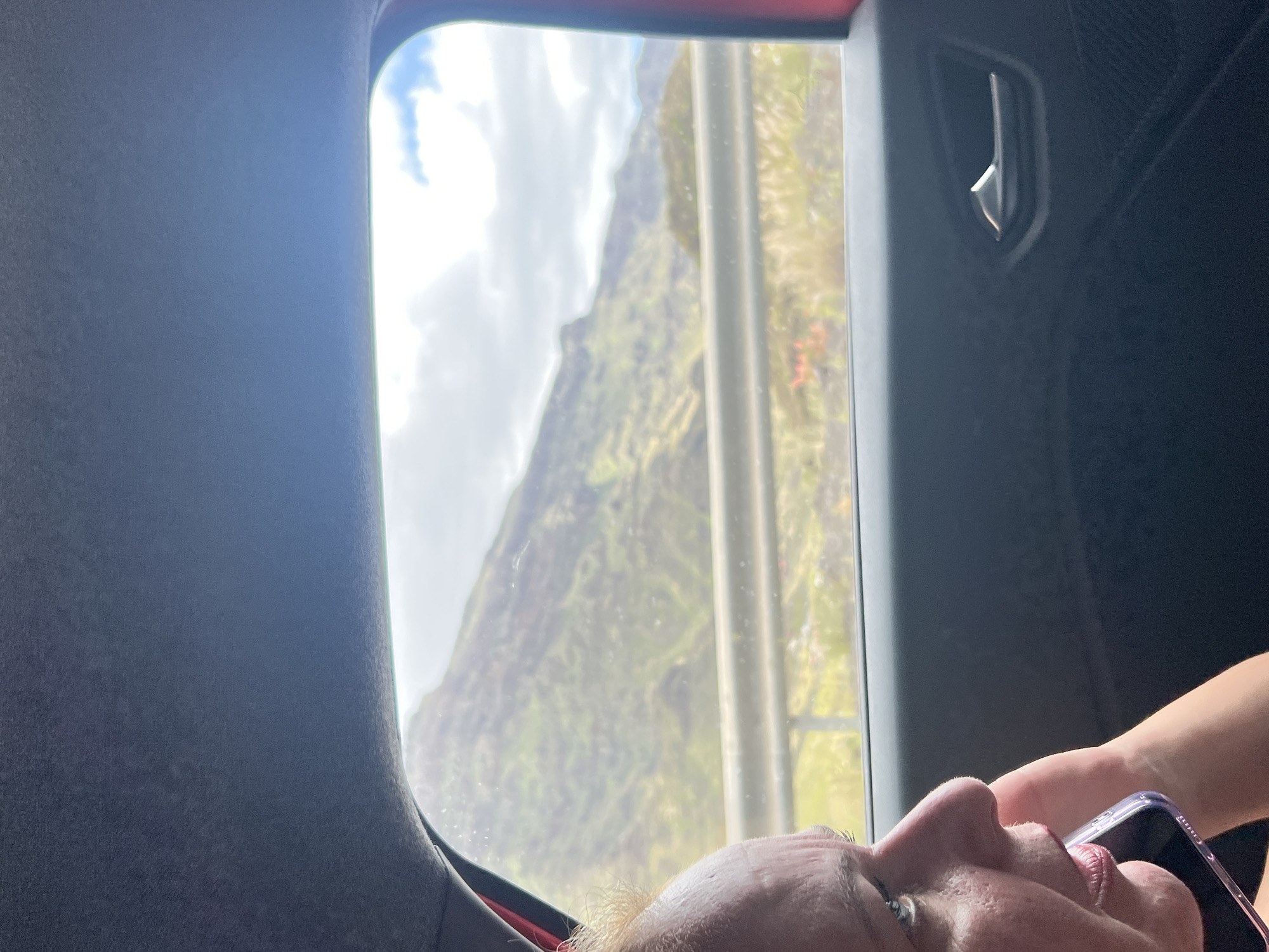 The image, taken from inside a vehicle, shows a person looking at a phone with a mountainous landscape visible through the window. The location is likely in Mogán, Spain, suggested by the arid, volcanic mountains characteristic of the Canary Islands. The window features a metal handle and a grey surround. The interior of the vehicle appears to be dark, and the person's face is visible in the lower portion of the frame.