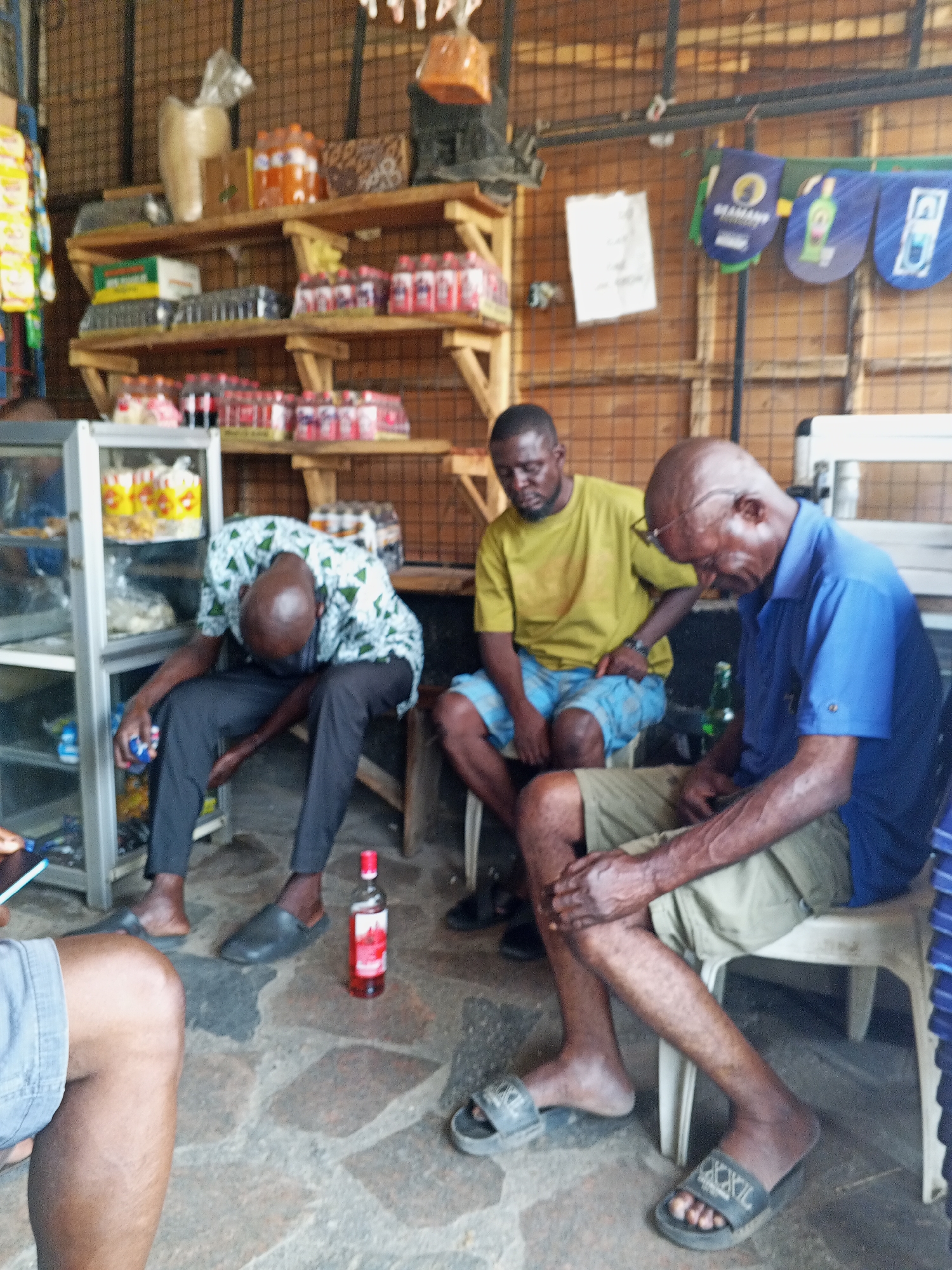 Three men are sitting on plastic chairs in a store in Zango-ndaji, Nigeria. The man on the left is wearing a patterned shirt and dark pants and appears to be looking down or reaching for something near the floor. A bottle of liquor sits on the floor near him. The man in the middle wears a yellow shirt and blue patterned shorts. The man on the right is wearing a blue shirt and khaki shorts. There is an open bottle of what appears to be liquor by him. The store has shelves stocked with various goods, including bottled drinks and snacks. The walls are made of wood. Other individuals can be partially seen in the image.