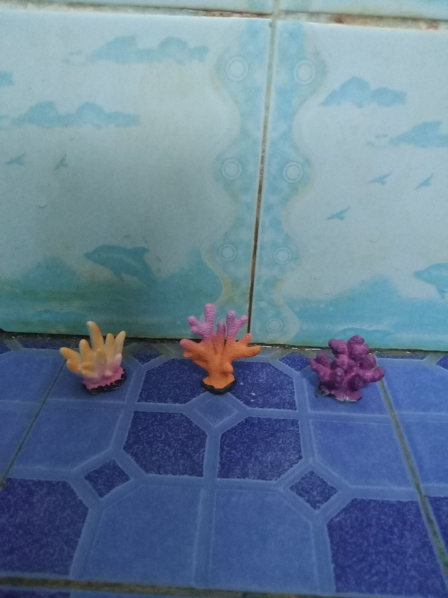 Three small, colorful artificial coral figurines are arranged on a blue tiled floor in front of a tiled wall. The wall is decorated with a mural depicting a marine scene, featuring clouds, birds, and a dolphin. The coral figurines are positioned from left to right: a yellow and pink one, an orange and pink one, and a purple one. The tiles on the wall have a light blue base with darker blue wave-like patterns and cloud shapes. The floor tiles are a dark blue with lighter blue hexagonal patterns. The scene appears to be a decorative display, possibly in a residential setting. The image was taken in the city of Kutosari, Indonesia.