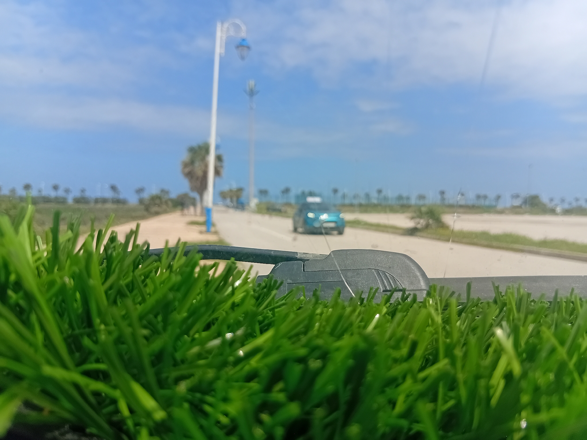 The image captures a vibrant, low-angle perspective looking over a patch of artificial green grass in the immediate foreground, which is in sharp focus. Behind the grass, a dark gray barrier or edge is partially visible.
The scene transitions to a blurred midground featuring a paved road or pathway. A light blue or turquoise car is seen driving on this road in the distance, heading away from the viewer. To the left of the road, a distinct white lamppost with a blue lantern-style fixture stands tall. Further along, another less distinct utility pole is visible.
The background is heavily blurred but reveals a landscape with scattered palm trees and low vegetation. In the far distance, a vast expanse of lighter blue, likely the sea, meets a clear, bright blue sky adorned with sparse, wispy white clouds.
No people are visibly present. The bright, clear lighting indicates it is daytime, likely midday or early afternoon, under sunny weather conditions. The setting, with palm trees, a coastal road, and the visible sea, is characteristic of a seaside town. The image evokes a sense of calm and openness. Given the location context of Martil, Morocco, this depiction aligns with its Mediterranean coastal environment. No visible text is present.