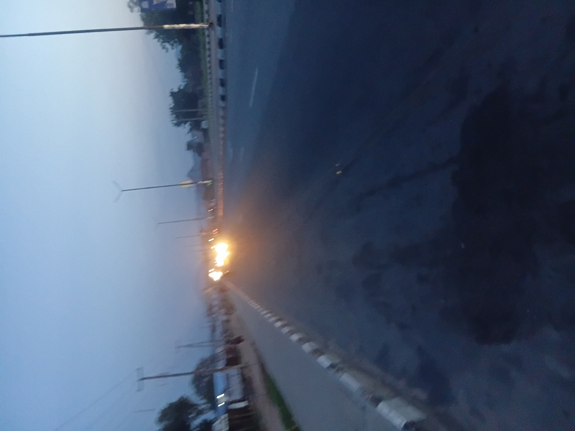 The image shows a road with streetlights at dusk or dawn. The sky is a pale blue, suggesting either early morning or late evening. Trees and some structures are visible on the left side of the road, with a few lights on in the buildings. The road itself is asphalt and appears to be a multi-lane highway with a white line marking the lanes. There are also white and black striped barriers along the side of the road. A few vehicles are present, with their headlights illuminated, indicating low visibility. The overall atmosphere is calm and quiet, characteristic of a transitional time of day. The scene is set in Jalingo, Nigeria. No text is visible in the image.