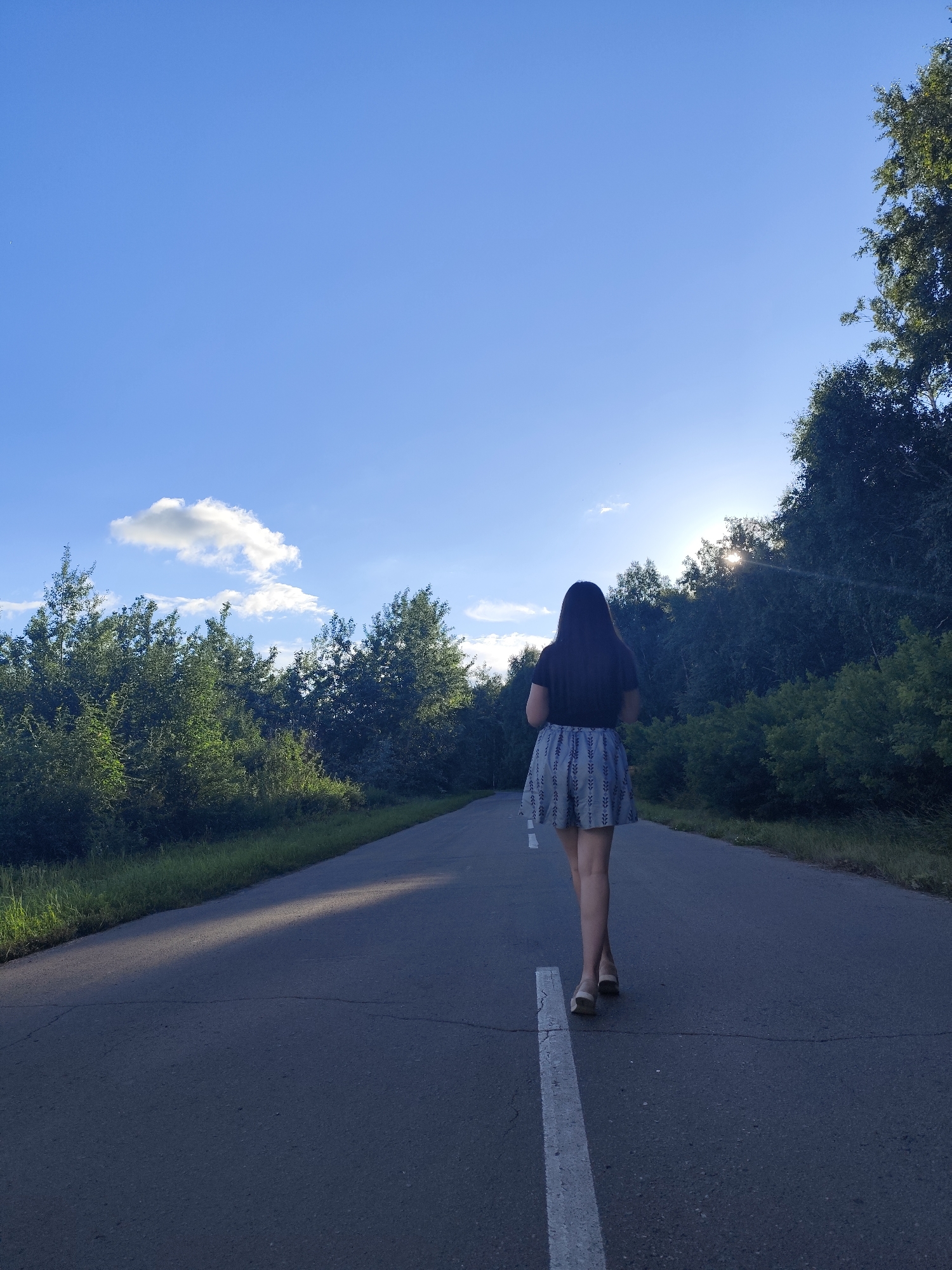 A full shot, from the rear, shows a young woman with long, dark hair walking away from the camera down a paved road. She is wearing a black t-shirt and a patterned skirt. The sun is low in the sky, casting long shadows across the road and illuminating the trees to the right of the scene. The road appears to be in a rural or semi-rural area, surrounded by dense green foliage. The sky is a clear blue with a few wispy white clouds.