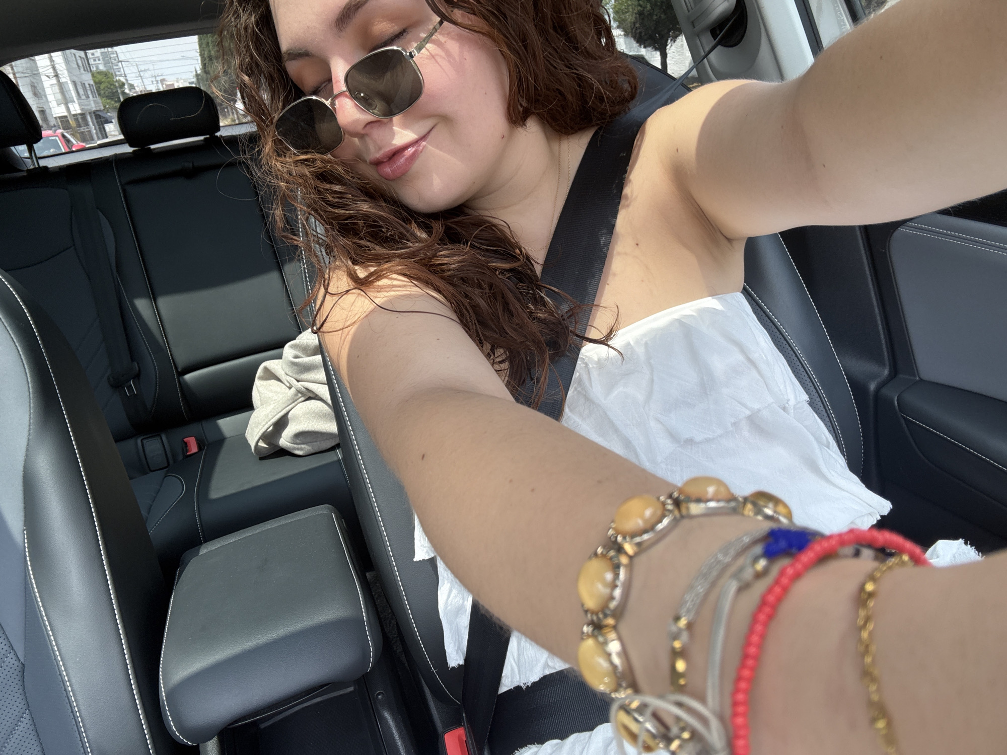 A person with curly brown hair and sunglasses is taking a selfie in a car. They are wearing a white top and a seatbelt. The car's interior is black leather with white stitching. The person is wearing several bracelets on their arm, including one with yellow stones and another with red beads. The back seat of the car is visible, and a grey garment is resting on it. The photograph was likely taken in the city of Puebla, Mexico, based on the buildings visible through the window.