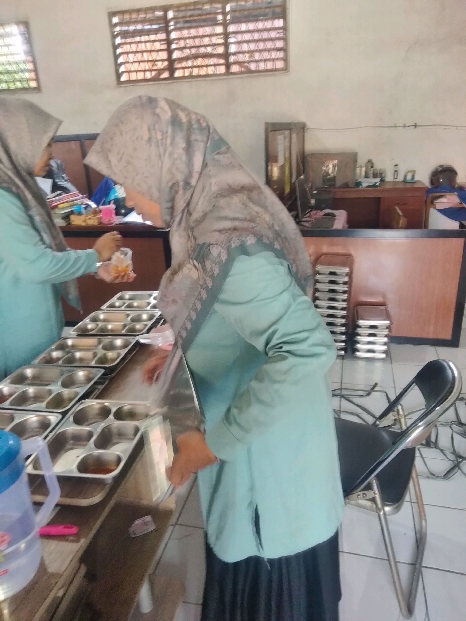 The image depicts an indoor scene, possibly a school cafeteria or serving area, located in Suka Sari, Indonesia. Two individuals wearing light blue uniforms and headscarves are present. The individual on the right is seen from the side, looking down and holding a clear plastic bag containing food. The individual on the left is partially visible, also in uniform. In the foreground, several stainless steel trays with multiple compartments are arranged on a counter. Some of these trays contain food. A large plastic jug with a blue lid and a pink object are also on the counter. In the background, there is a wooden counter and a dark brown partition. Stacked stainless steel containers are visible to the right of the partition, along with a black folding chair. The environment appears to be a functional space with tiled flooring and a window with louvers in the upper left background.