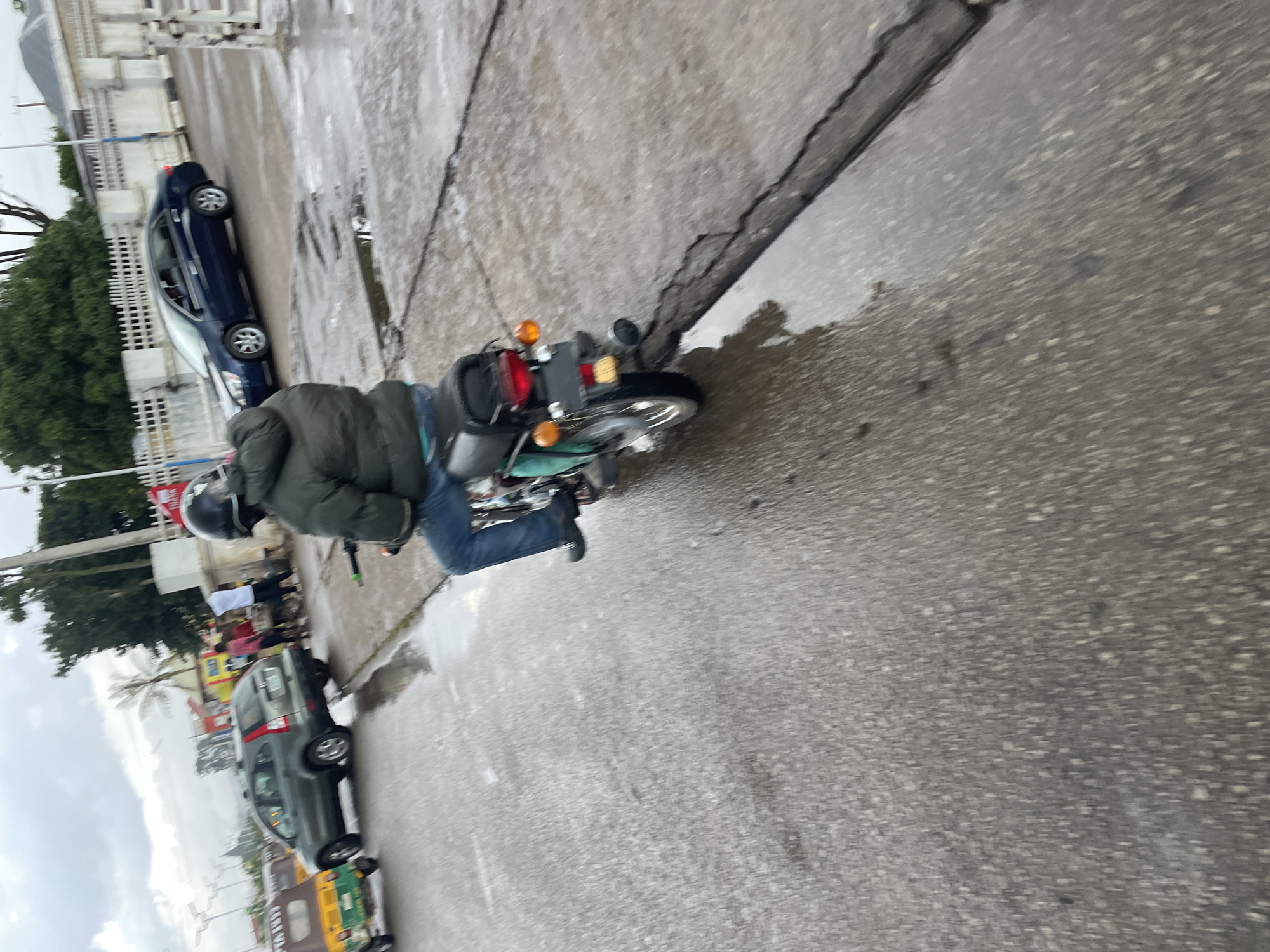 The image shows a person riding a motorcycle on a wet, cracked asphalt road in Barakin Lerawi, Nigeria. The rider is wearing a dark puffer jacket and blue jeans, with a black helmet. The motorcycle is green and appears to be an older model with a black seat and a red taillight. In the background, several cars are visible, including a dark blue sedan and a grey SUV. There are also a few people standing on the side of the road, near what appears to be a building with white fencing. The sky is overcast, suggesting it may have recently rained or is about to. The overall scene depicts a typical street in a Nigerian city, with active traffic and pedestrians.