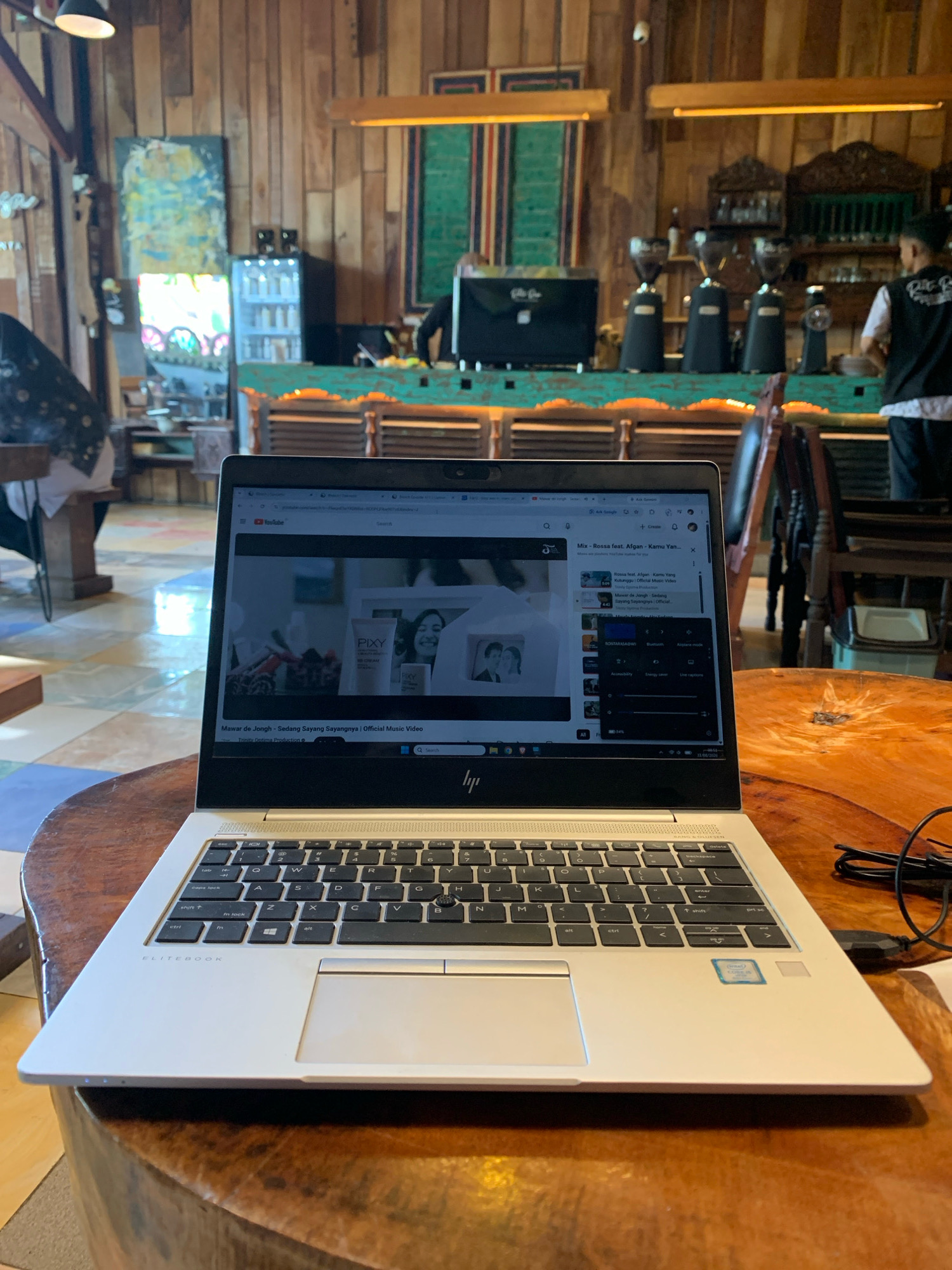 In Lhokseumawe, Indonesia, a laptop is positioned on a round wooden table in what appears to be a cafe or a similar establishment with a rustic, wooden interior. The laptop screen displays a YouTube video. The video features an advertisement for "PIXY BB CREAM" with a woman's face visible, and the song title "Mawar de Jongh - Sedang Sayang Sayangnya | Official Music Video" is displayed. The laptop itself is a silver HP Elitebook. To the right of the laptop, a black USB cable is plugged into the side. The background reveals a bar area with coffee grinders and shelves stocked with bottles. In the distance, individuals are seen, one of whom is wearing a dark vest over a white shirt. The lighting suggests it could be daytime, with artificial lights also present.