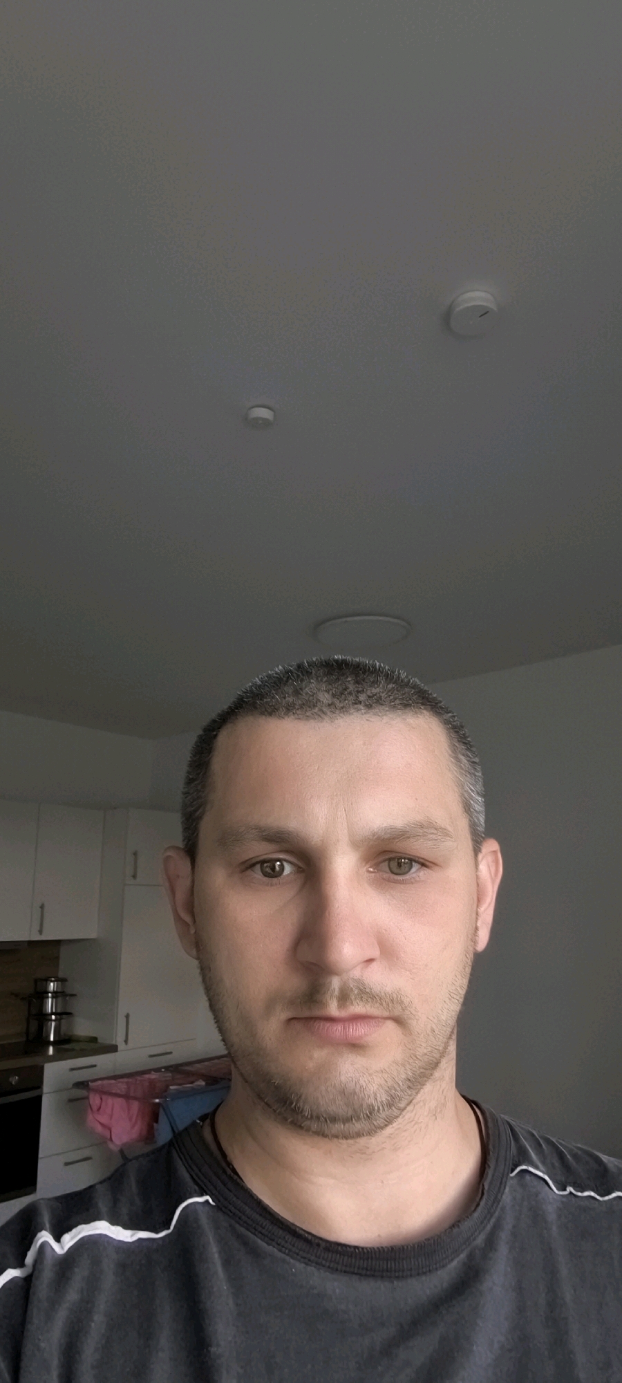 The image is a close-up, frontal portrait of a light-skinned man with short, dark hair and stubble on his face. He is wearing a dark t-shirt with a white striped detail around the collar. His eyes are open, and he is looking directly at the camera. The background appears to be an indoor setting, possibly a kitchen, with white cabinets visible to the left. Two circular ceiling fixtures are also present in the upper portion of the image. The man is approximately in his late 20s to early 30s. The location is not specified within the image itself.