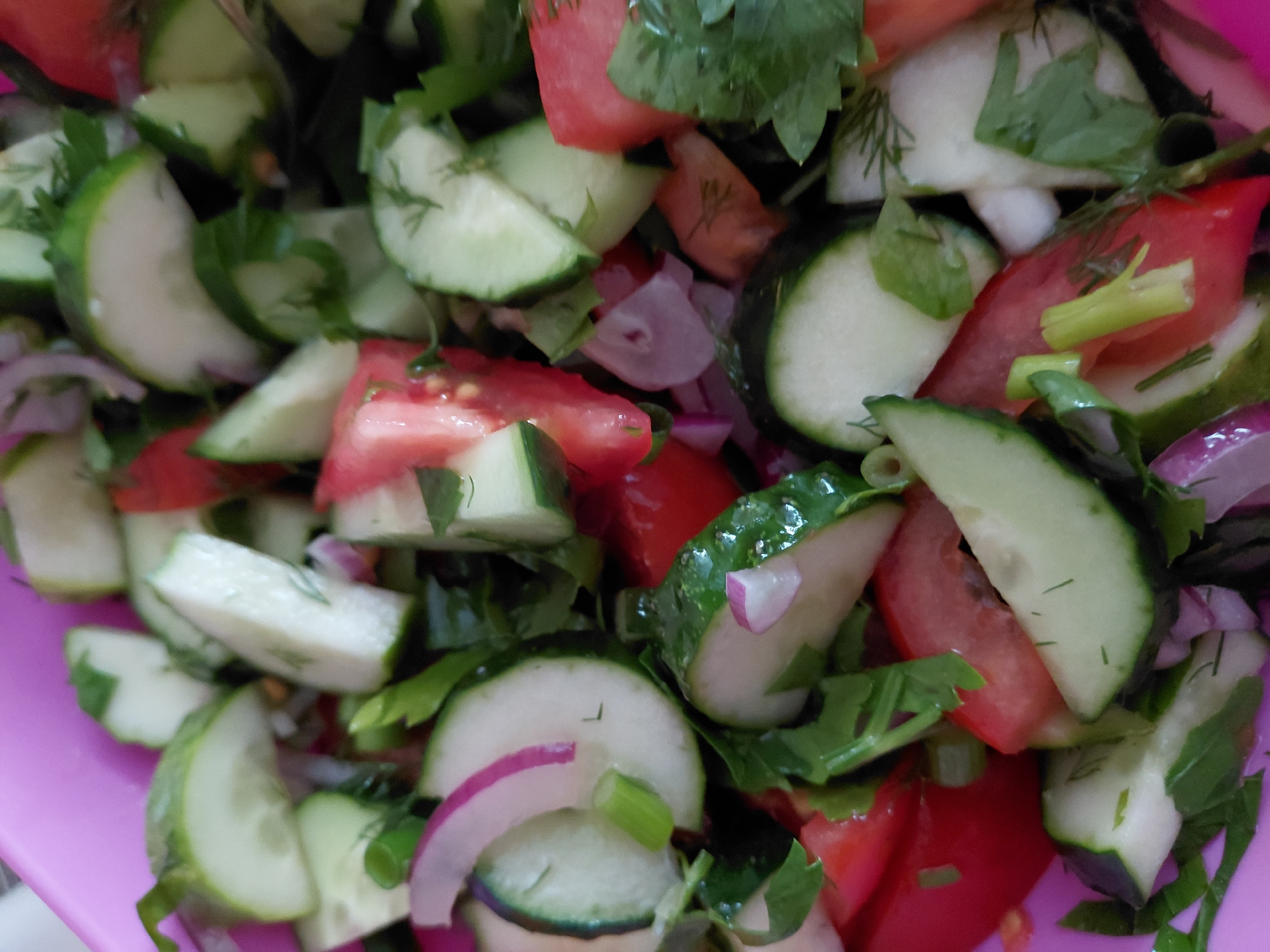 A close-up, overhead shot shows a chopped salad in a pink bowl. The salad consists of sliced cucumbers, diced tomatoes, and thinly sliced red onions, mixed with chopped green herbs, possibly dill and parsley. The ingredients are jumbled together, with no discernible pattern. The salad appears fresh and vibrant, with the colors of the vegetables contrasting against each other. The pink bowl is visible at the edges of the frame. The scene is well-lit, likely by natural light, highlighting the textures and details of the ingredients. There are no people visible in the image, and no specific location in Bila Tserkva, Ukraine can be identified.
