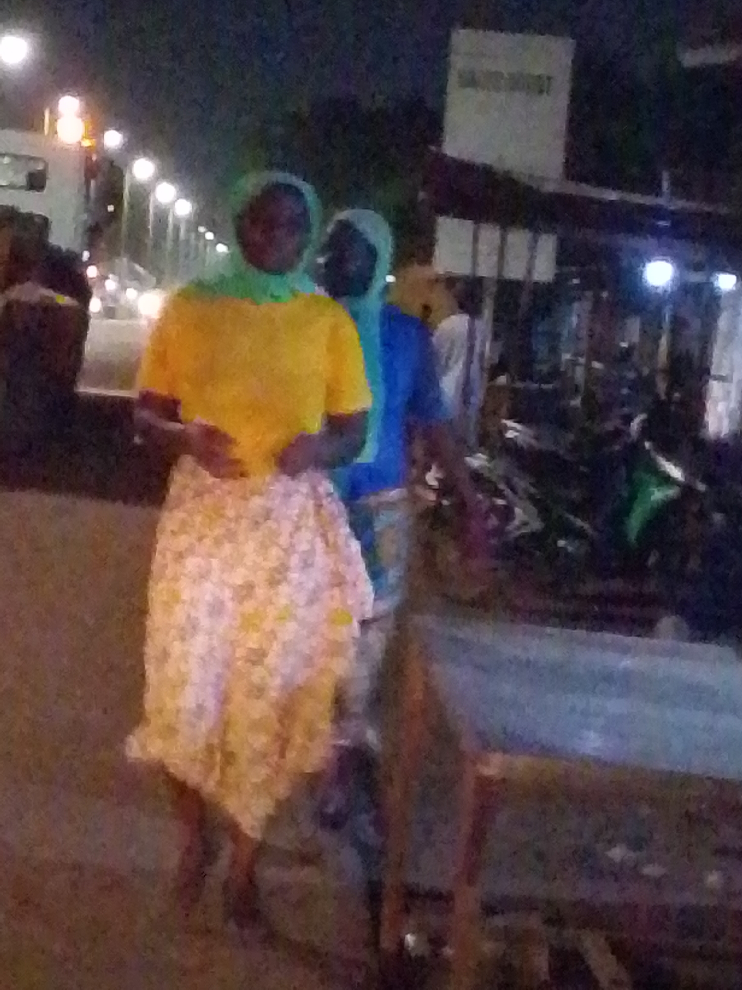 The image shows two people standing on a sidewalk at night in Tamale, Ghana. The person in the foreground is wearing a yellow shirt, a floral skirt, and a head covering. The second person is wearing a blue shirt and a head covering. Behind them, there is a busy street with cars and streetlights, and a building with a sign. To the right of the people, there is a table and what appears to be a motorcycle parked in front of a shop.
