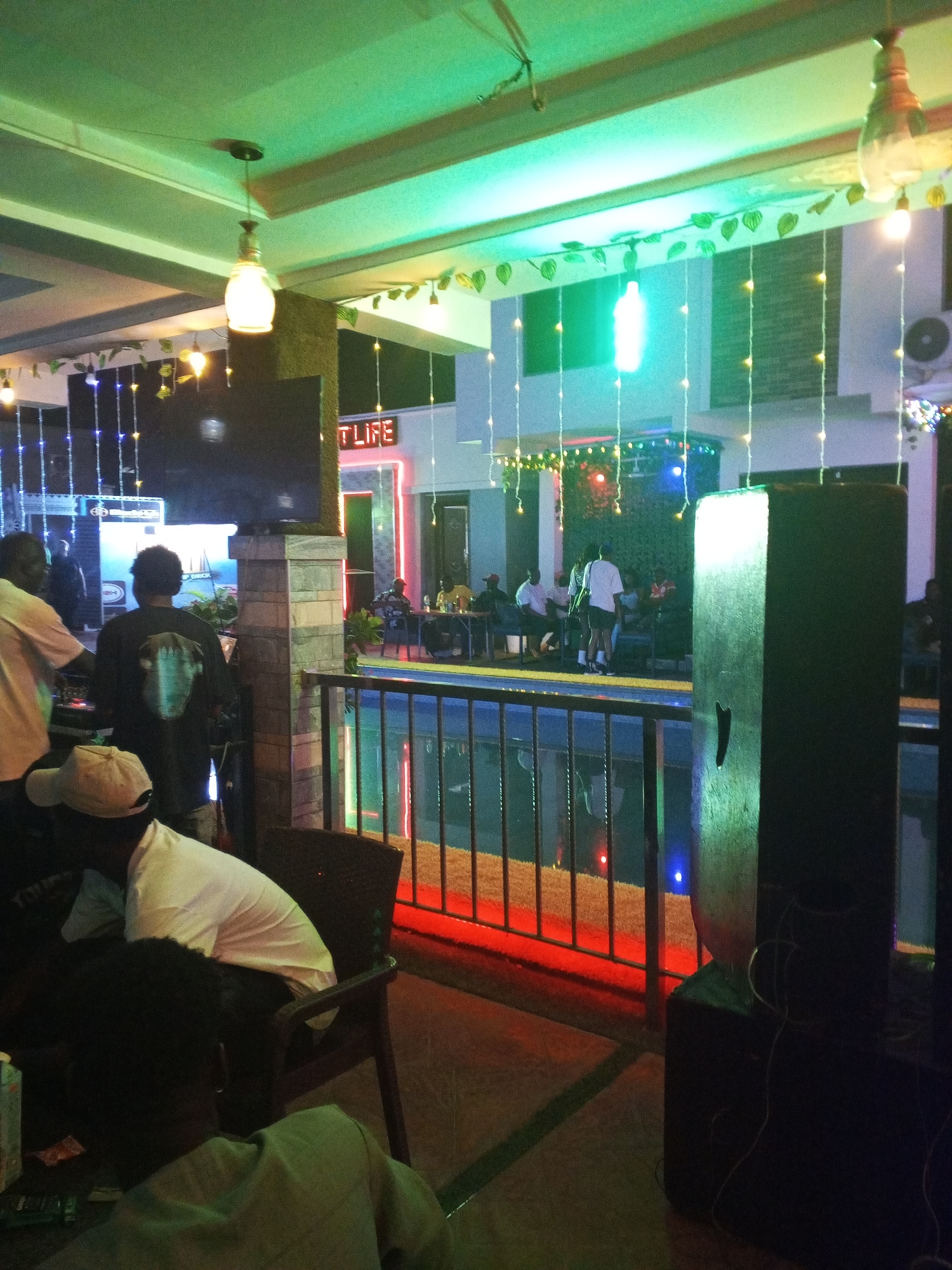 The image depicts an indoor/outdoor event, likely at a bar or lounge in Lokoja, Nigeria. A band is performing on a stage that appears to be adjacent to a pool. The setting is lit with colored lights and string lights. The phrase "T LIFE" is visible in neon signage. There are several people seated at tables, some watching the band and others seemingly socializing. A large speaker is prominent in the foreground of the image.