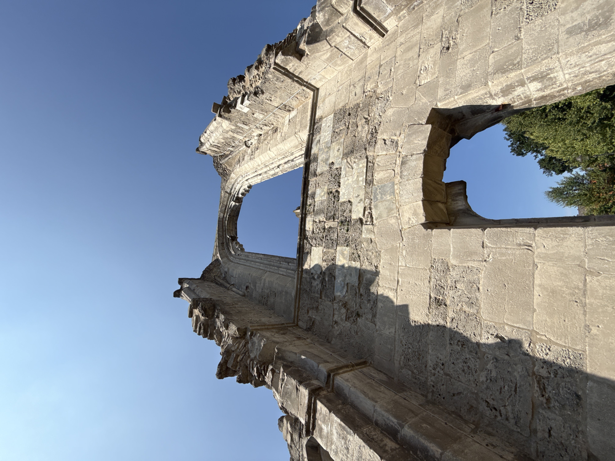 The image shows the ruins of a Gothic-style church, likely the Walkenried Abbey, located in Walkenried, Germany. The photograph is taken from a low angle, looking up at the stone ruins against a clear blue sky. The focus is on the weathered stone structures, including arched openings and crumbling walls, which are constructed from large, rough-cut blocks. Sunlight casts shadows across the textured surfaces of the stone. In the lower right portion of the frame, through an archway, a section of green trees is visible against the blue sky. The overall impression is one of historical decay and enduring architecture.