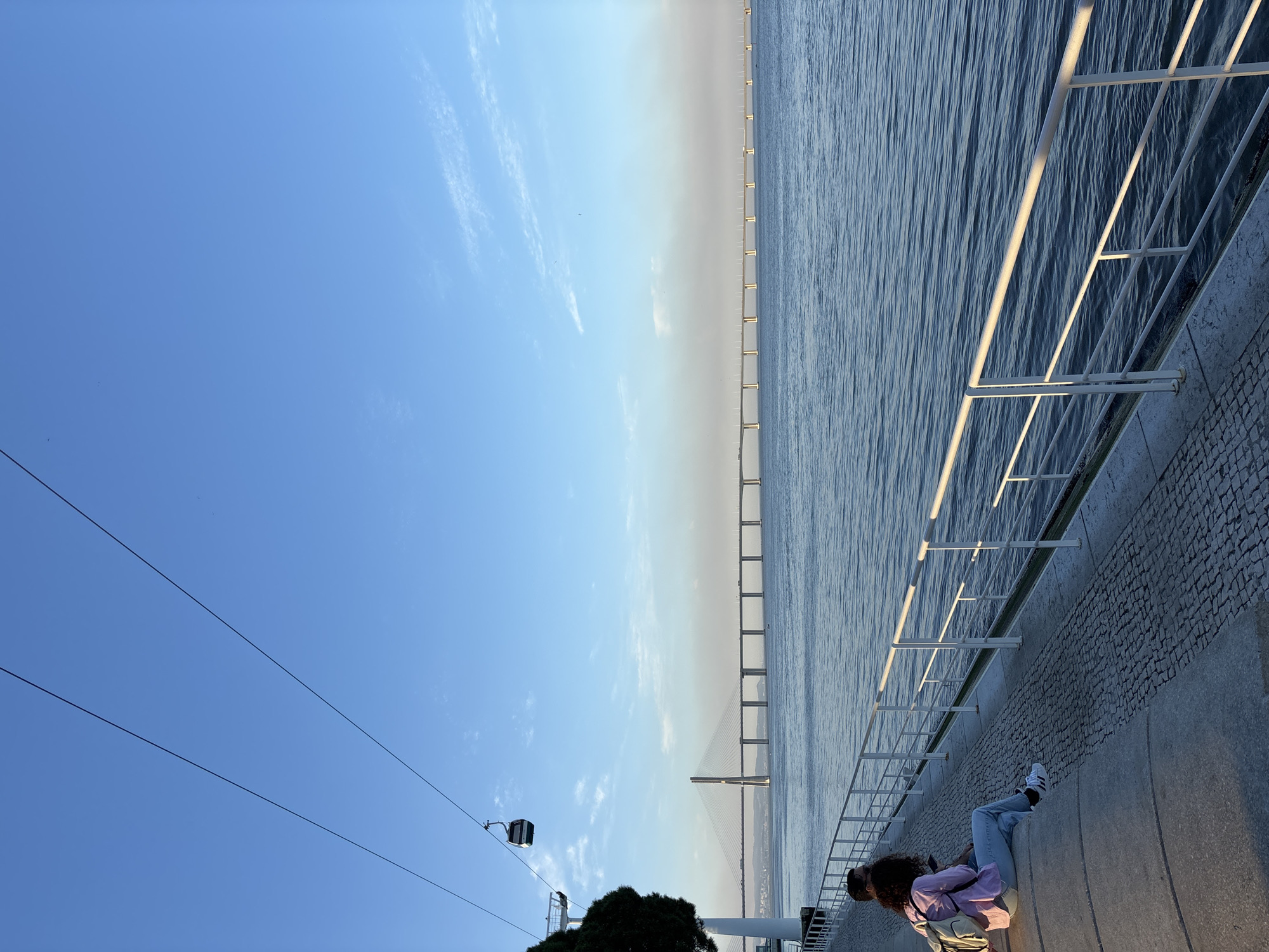 The image captures a sunny day in Lisbon, Portugal. The Vasco da Gama Bridge stretches across the Tagus River in the background. A cable car line is visible in the left foreground, with one of the cable cars suspended on the line. A person sits on a low wall along the riverfront. White railings line the walkway beside the water, with lights attached below. The sky is clear blue, with some light clouds present. The river's surface is slightly rippled.