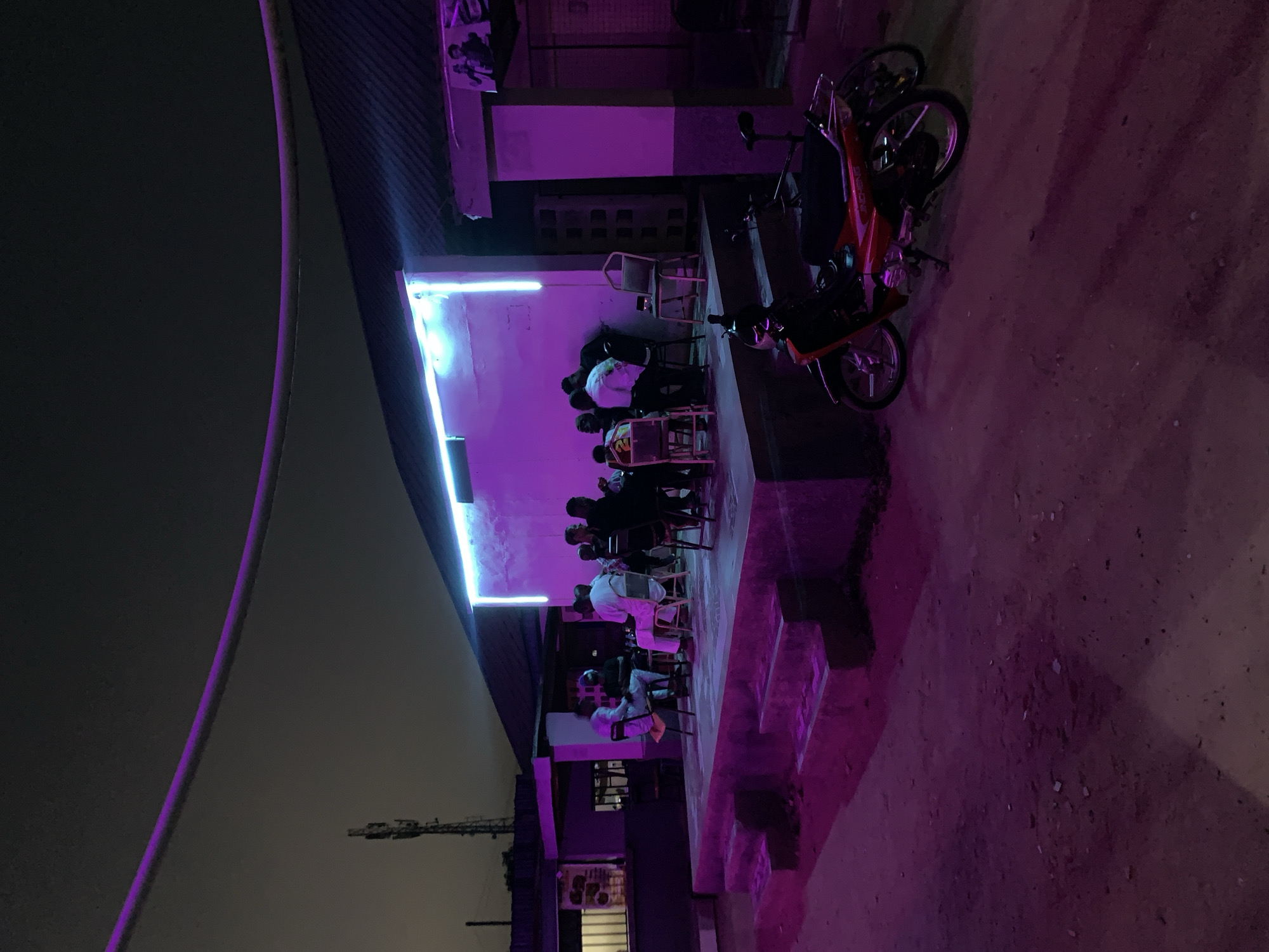 The image depicts an outdoor scene in New Stadium, Nigeria, at night. The area is illuminated by a purple hue, possibly from neon lights. A group of people are seated on chairs, congregating under a roof that has rectangular neon lights. A red motorcycle is parked nearby. The ground appears to be dirt or gravel. A tall structure, possibly a mast, is visible in the background against a dark sky.