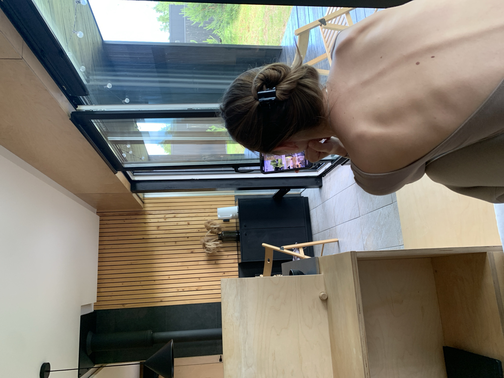 A person's back is visible as they face a tablet device showing a video call, in a modern room with wooden paneling. The room appears to be a home, possibly in the city of Мяжанскі сельскі Савет, Belarus, given the architecture and outdoor scenery. A large glass sliding door opens to a view of greenery and a structure, suggesting a rural or semi-rural setting. The person is wearing a light-colored top and has their hair pulled back in a bun with a clip. The tablet is held in their hands, and they are looking intently at the screen. The room features a dark cabinet, a folding chair, and some decorative items. The lighting suggests daytime.