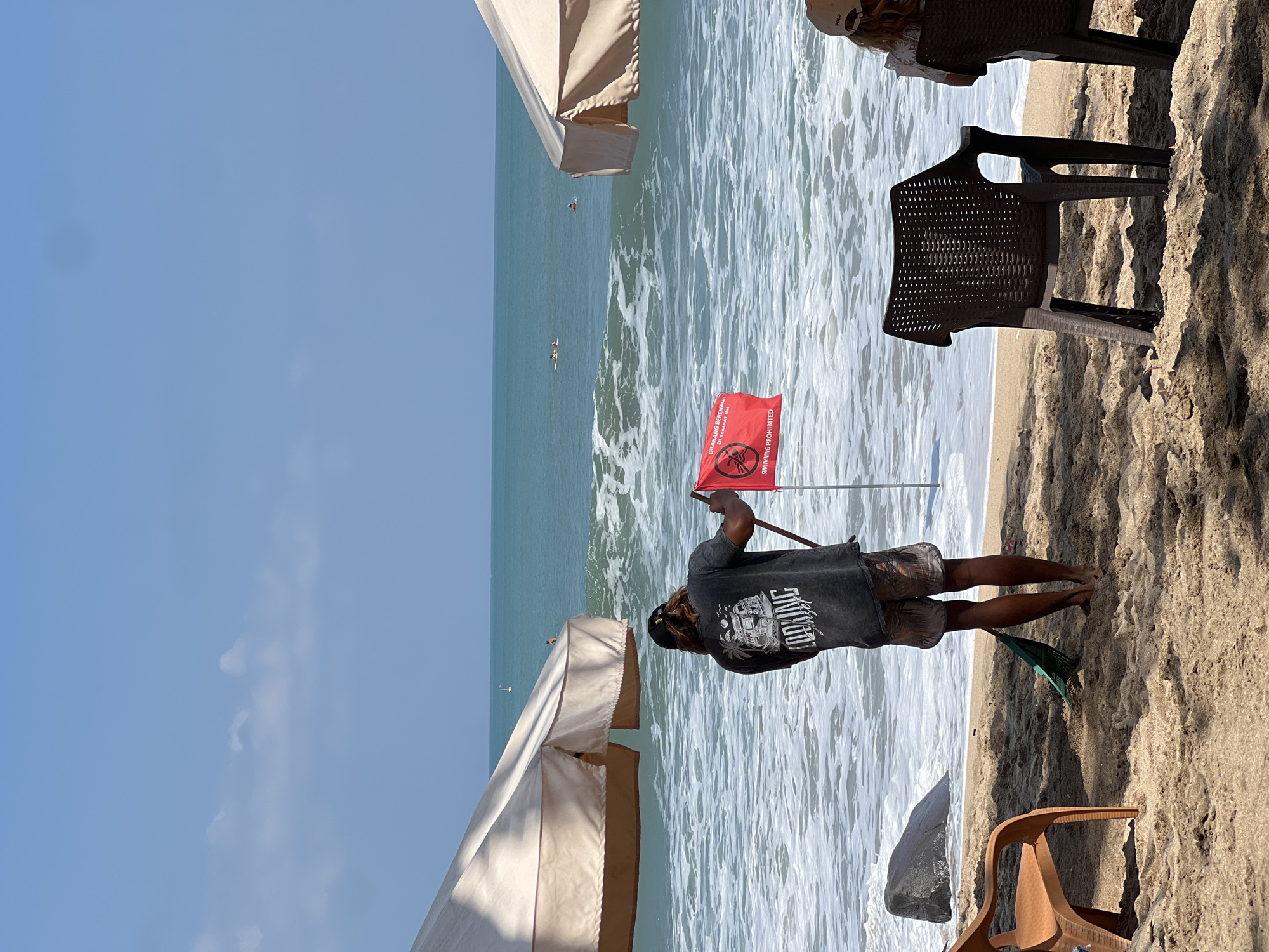 In Kuta, Indonesia, on a sandy beach, a person stands near the water's edge, holding a red flag with a white symbol and text. The flag reads "DILARANG BERENANG DI TEMPAT INI" and "SWIMMING PROHIBITED." The person is wearing a dark t-shirt with graphics and patterned shorts, and is holding a green rake. In the background, the ocean has moderate waves with white foam. Two white beach umbrellas are visible on either side of the frame, casting shadows on the sand. Several plastic chairs are placed on the sand. Two individuals are in the ocean, one on a surfboard and another swimming. The sky is clear blue with a few scattered clouds.