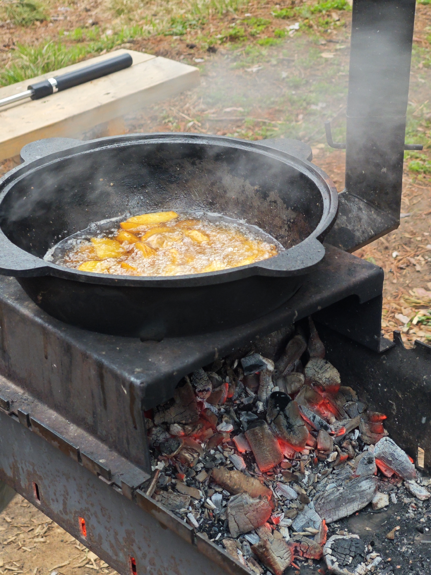 This outdoor scene, set in Мишкино, Russia, captures food being cooked in a large, black cast-iron pot, commonly known as a kazan. The pot is placed directly over a metal grill or brazier (mangala) filled with glowing red and grey charcoal embers, indicating active cooking. Inside the pot, pieces of what appear to be potatoes are deep-frying or pan-frying in bubbling oil, with steam or smoke visibly rising from the hot food and oil.
The grill itself is a sturdy metal structure, showing signs of use and heat, with some visible rust and wear. To the right of the pot, a vertical chimney or smoke stack is integrated into the grill, directing smoke upwards. In the background, a wooden board lies on the ground, and on it rests a long, black and silver metal tool, possibly a skewer or a handle for a cooking utensil. The ground consists of dirt and patches of green and dry grass, suggesting a natural outdoor environment like a backyard or a picnic area. The lighting indicates it is daytime. No people are visible, and there is no visible text in the image.