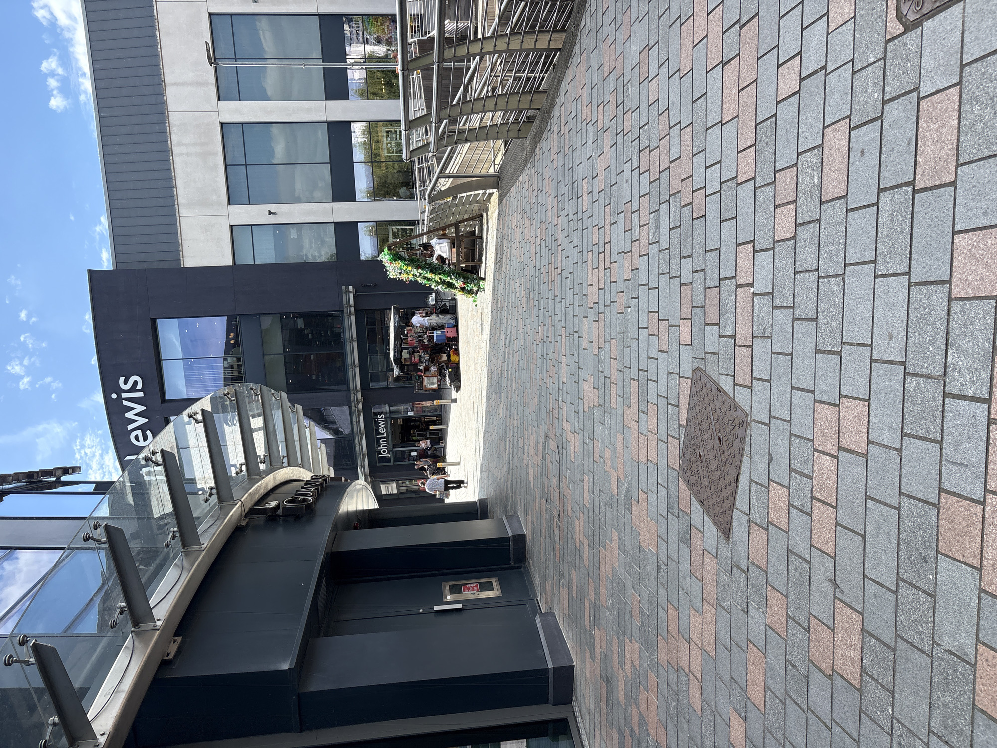 The image shows a street scene with a John Lewis department store visible. The store has a dark facade with large windows reflecting the sky and surrounding buildings. A glass and metal entrance canopy curves over a doorway. The street itself is paved with rectangular cobblestones in shades of gray and pink. In the middle ground, people are visible near the entrance of the store. Some are standing, while others appear to be browsing. A decorative, possibly festive, green structure with lights is situated on the right side of the entrance. The sky above is blue with scattered white clouds. The time of day appears to be daytime, and the weather seems fair. The overall setting is an urban environment, likely a shopping district. Visible text includes "John Lewis" on the store's signage.