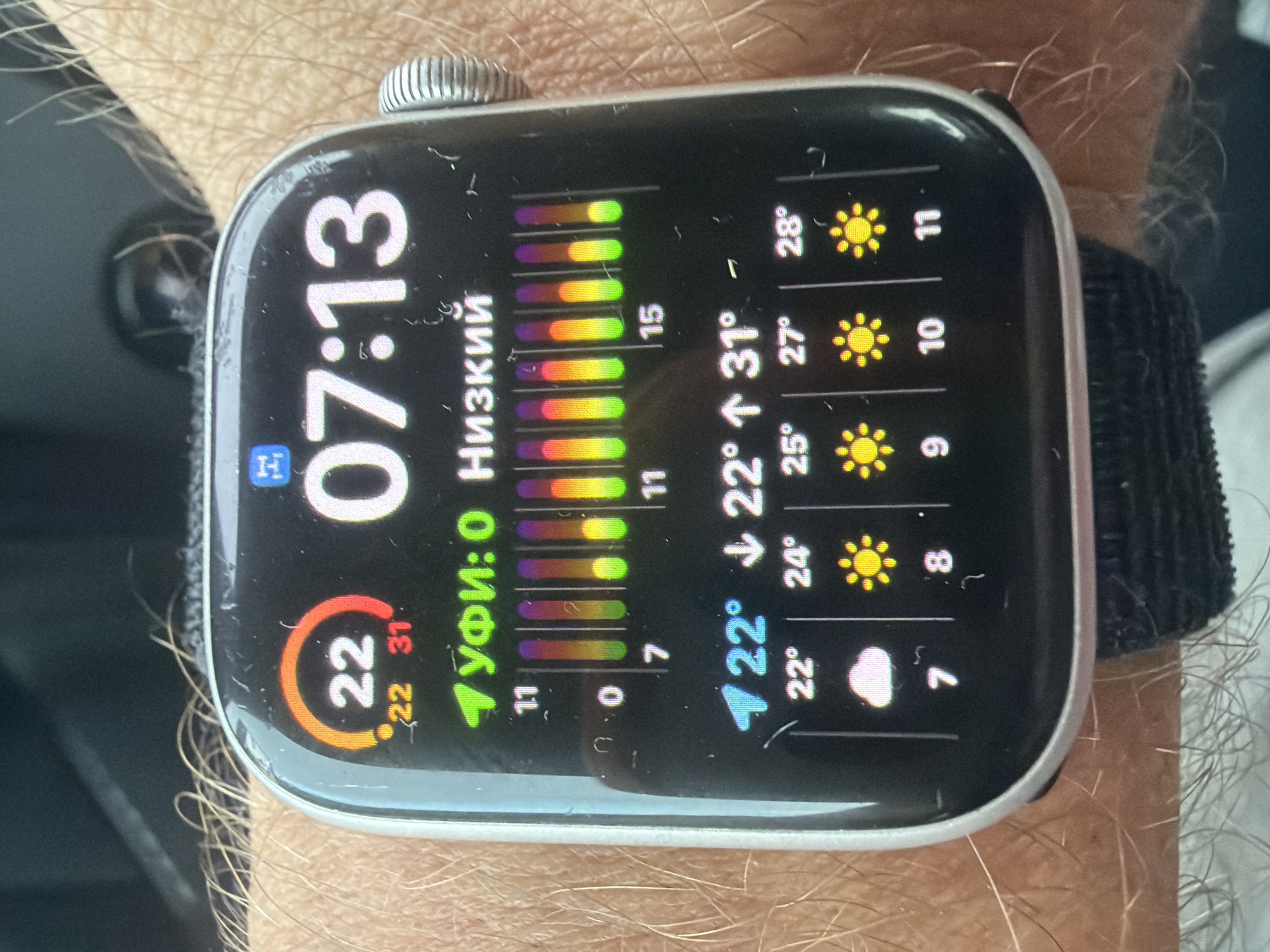 The image displays a close-up of a smartwatch on a person's wrist. The smartwatch screen shows the time as 07:13. It also displays weather information, including a temperature of 22 degrees, with a forecast for 22 and 31 degrees. Below this, there is a graphic with vertical bars and labels "УФИ:0 Низкий" which likely indicates UV index and air quality. Further down, there are hourly weather forecasts with temperatures and weather icons: 22° with clouds at 7, 24° with sun at 8, 25° with sun at 9, 27° with sun at 10, and 28° with sun at 11. The background is blurred, showing the person's arm with hair and what appears to be fabric or a surface. The context of "Wildwood, United States" does not appear to be directly visible or inferable from the image content itself.