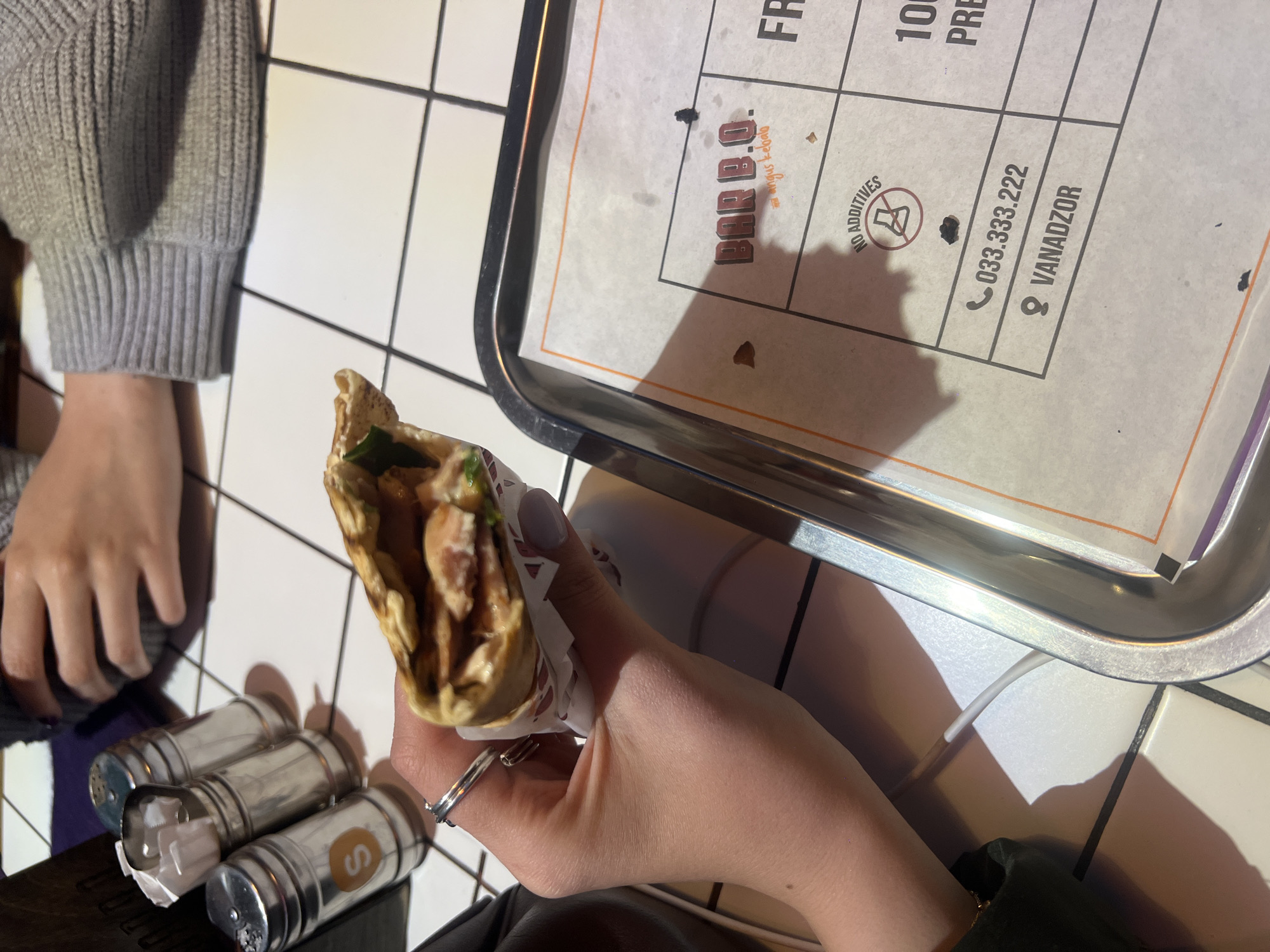 The photograph captures a still life scene set on a white tiled surface, likely a table, at what appears to be a restaurant. A person with a gray sweater sleeve visible on the left is seated at the table. Another person is holding a wrap or kebab in their right hand, the filling seemingly consisting of meat and greens. The person's fingers are adorned with a ring on their ring finger, and their nails are painted a light shade. A tray with a paper insert bearing the name "BAR.B.Q." is placed near the wrap. The paper also has the text "NO ADDITIVES" and a phone number, "033.333.222" followed by the location "VANADZOR". Three silver salt shakers are positioned near the left side of the frame. Given the restaurant's location in Vanadzor, Armenia, the city of Yerevan is not directly depicted in the photo.