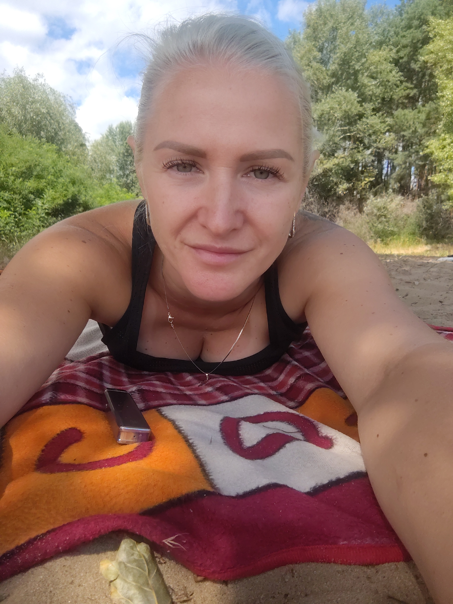 A blonde woman lies on a blanket on a sandy shore, with trees and a cloudy sky in the background. She is looking at the camera with a slight smile. Her hair is pulled back, and she is wearing a black tank top and a necklace. A cell phone and a leaf are on the blanket. The image appears to have been taken outdoors in a natural setting.