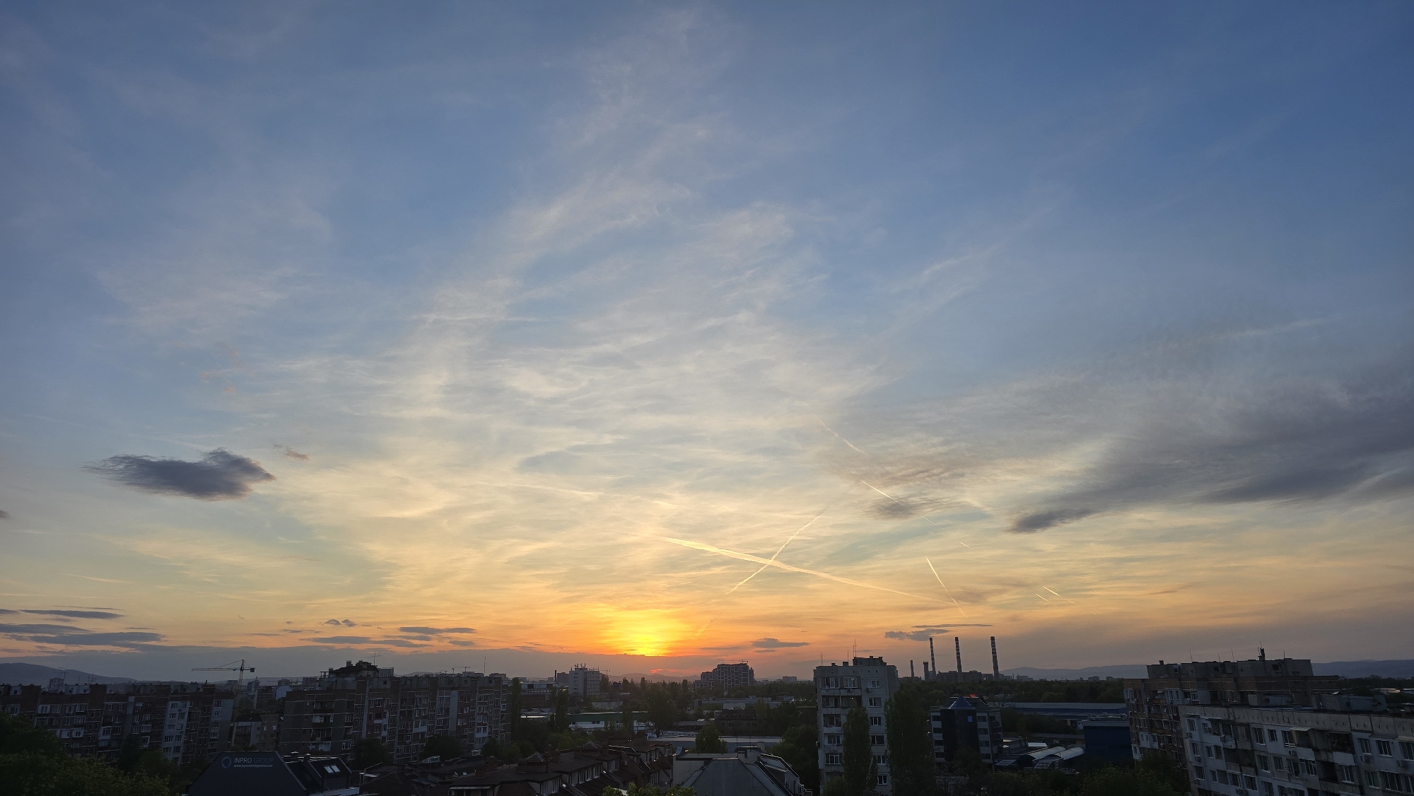 The image captures a vibrant sunset over an urban landscape in Sofia, Bulgaria, viewed from an elevated position. The sky occupies the majority of the frame, displaying a dramatic blend of colors. The setting sun, a bright orange and yellow orb, is situated low on the horizon, slightly left of the center, casting a warm glow across the lower cloud formations. Above, the sky transitions through shades of light blue, grey, and soft purple. Numerous wispy clouds, characteristic of cirrus, are spread across the sky, intersected by several distinct white contrails from aircraft. A more prominent, darker grey cloud is visible in the upper-left portion of the sky.
Below the horizon, the city skyline features a dense arrangement of multi-story residential buildings, primarily in tones of grey and brown, interspersed with green trees. In the distance, a hazy outline of mountains or hills stretches across the background. To the right, several tall industrial chimneys or smokestacks rise into the sky, suggesting an industrial presence beyond the residential areas. A construction crane is visible in the mid-ground, positioned among the buildings on the left side. On a building in the lower-left, white text against a dark facade reads "INPRO GROUP" and "www.inprogroup.com". The scene conveys the tranquil atmosphere of dusk over a bustling city.