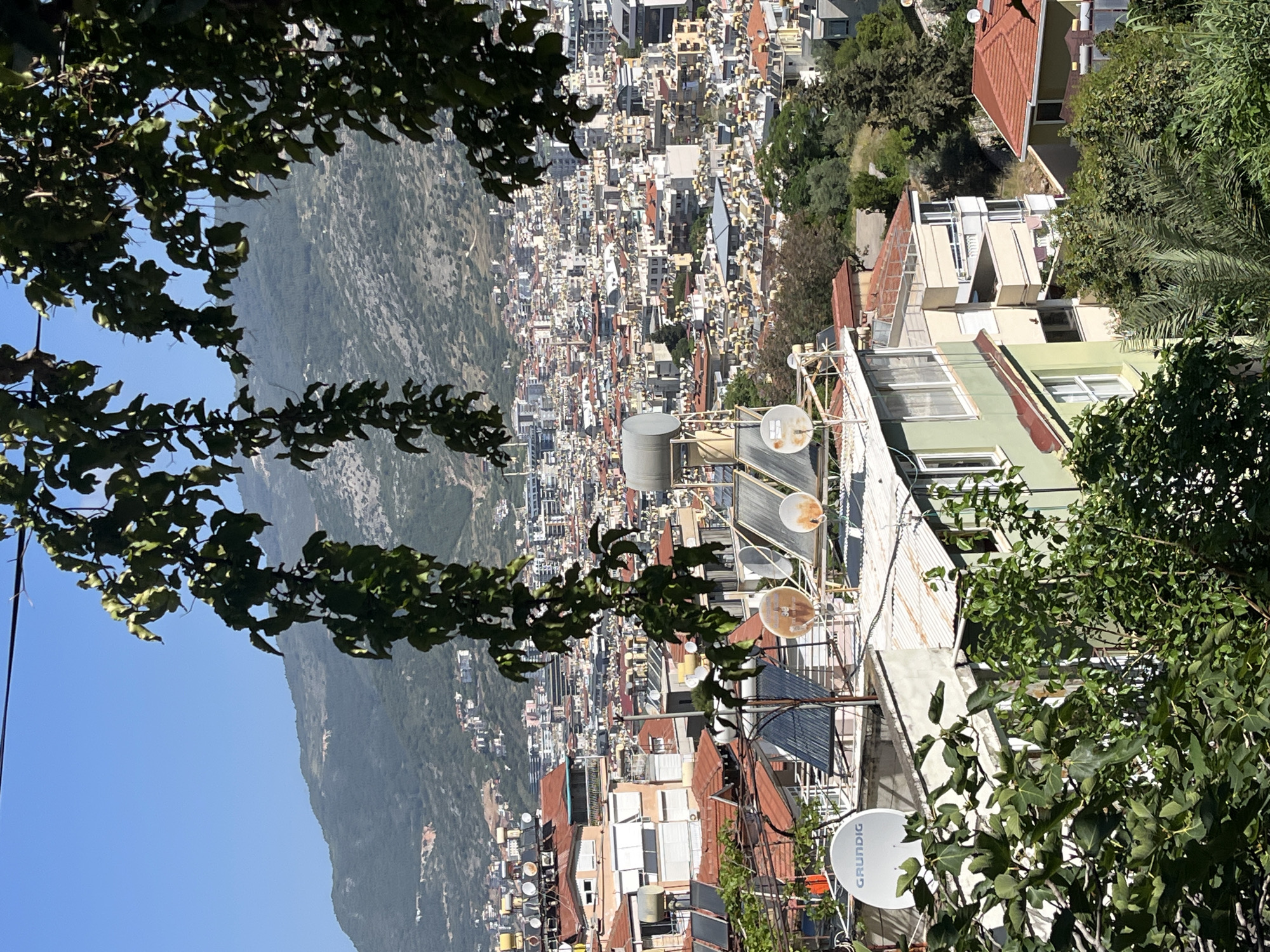 This photograph shows a high-angle, slightly rotated view of part of Alanya, Turkey. The cityscape, characterized by dense, mixed-use buildings, stretches toward a mountain range visible in the background. Patches of green vegetation are interspersed within the urban landscape. In the foreground, the view is partially obstructed by tree branches. Satellite dishes and rooftop water tanks are visible on the rooftops. The sky is clear.