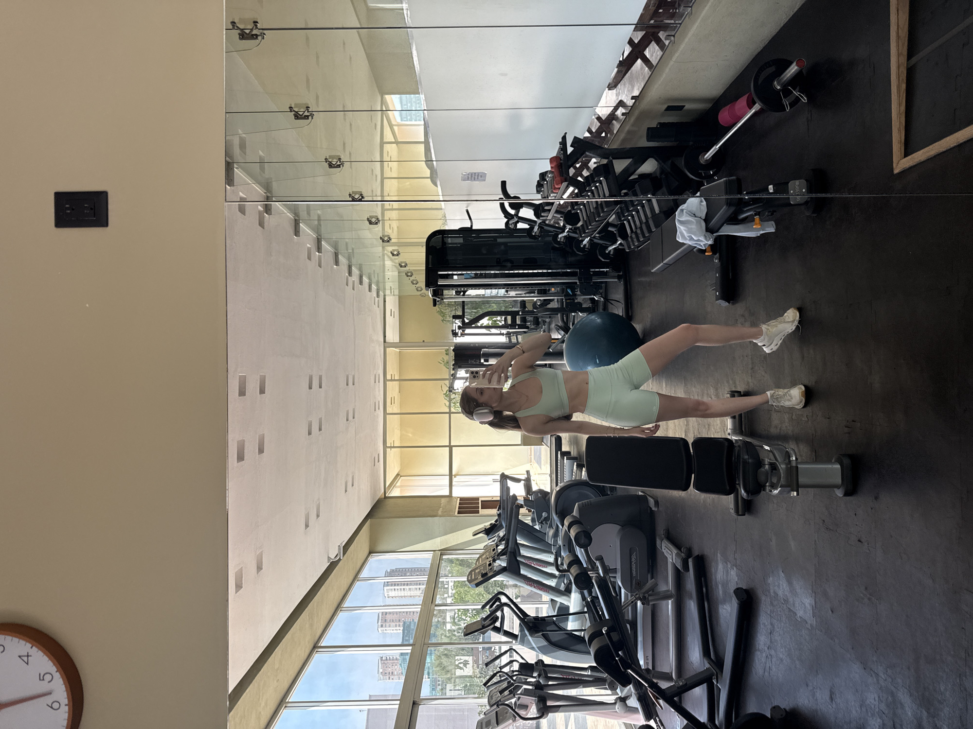 A woman in light green athletic wear and white headphones stands in a gym, holding a smartphone and facing a mirror that reflects the gym's interior, including treadmills, weight machines, and a blue exercise ball. The gym features a wall of windows revealing a cityscape, likely in Mexico City, Mexico, given the urban density and architectural style visible. The flooring is dark, possibly rubber, and the walls are light-colored, with a clock visible on the lower left. An electrical outlet is mounted on the wall next to the mirror.