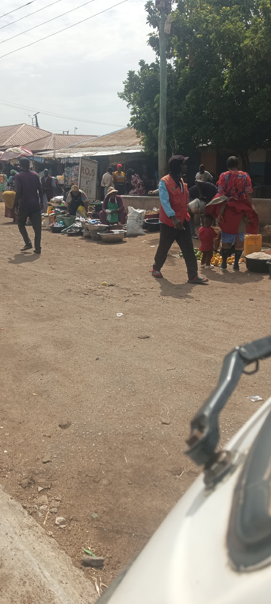 The image depicts a bustling outdoor market scene in Bukuru, Nigeria. Several people are present, including vendors and customers of different ages. A man in an orange vest and a black face mask is walking in the center, while a child in red stands nearby. Women are seen sitting by their wares displayed in bowls and on the ground.
The market is set on a dirt road, with simple structures providing shelter for some vendors. Various goods, including produce and other items, are displayed for sale. A power pole stands tall amidst the market activity. The foreground shows a portion of what appears to be a motorcycle.