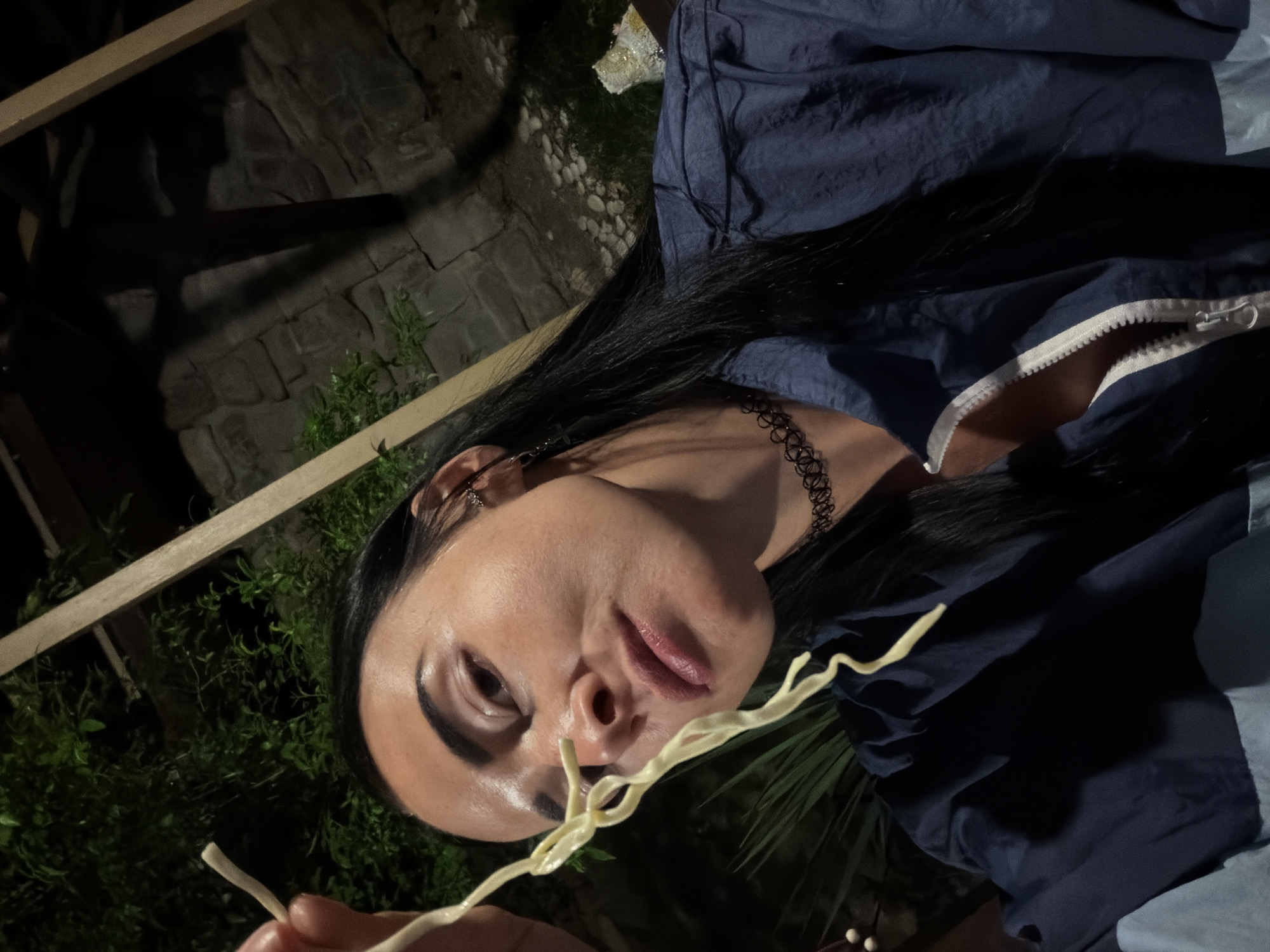 A woman with long, dark hair and a choker necklace is shown in a close-up shot, holding a strand of pasta up to her face. She is wearing a dark blue jacket. The background appears to be an outdoor setting at night, with stone walls and foliage visible. There is no indication of the specific location in Sochi, Russia.