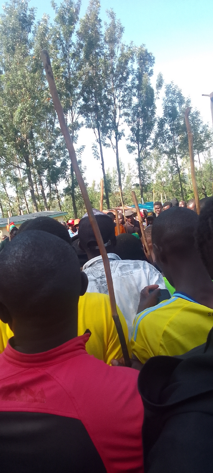 The image shows a group of people gathered in a forested area in Isukha ICHINA, Kenya (latitude: 0.351, longitude: 34.782). The individuals are mostly men, dressed in casual clothing. Some are wearing yellow, red and black shirts. A few people are carrying sticks or walking sticks. The group appears to be in a celebratory mood, with some smiling and laughing. The background shows a dense forest with tall trees and a blue sky. The scene is likely a social gathering or event, with a focus on the group of people at the center of the image.