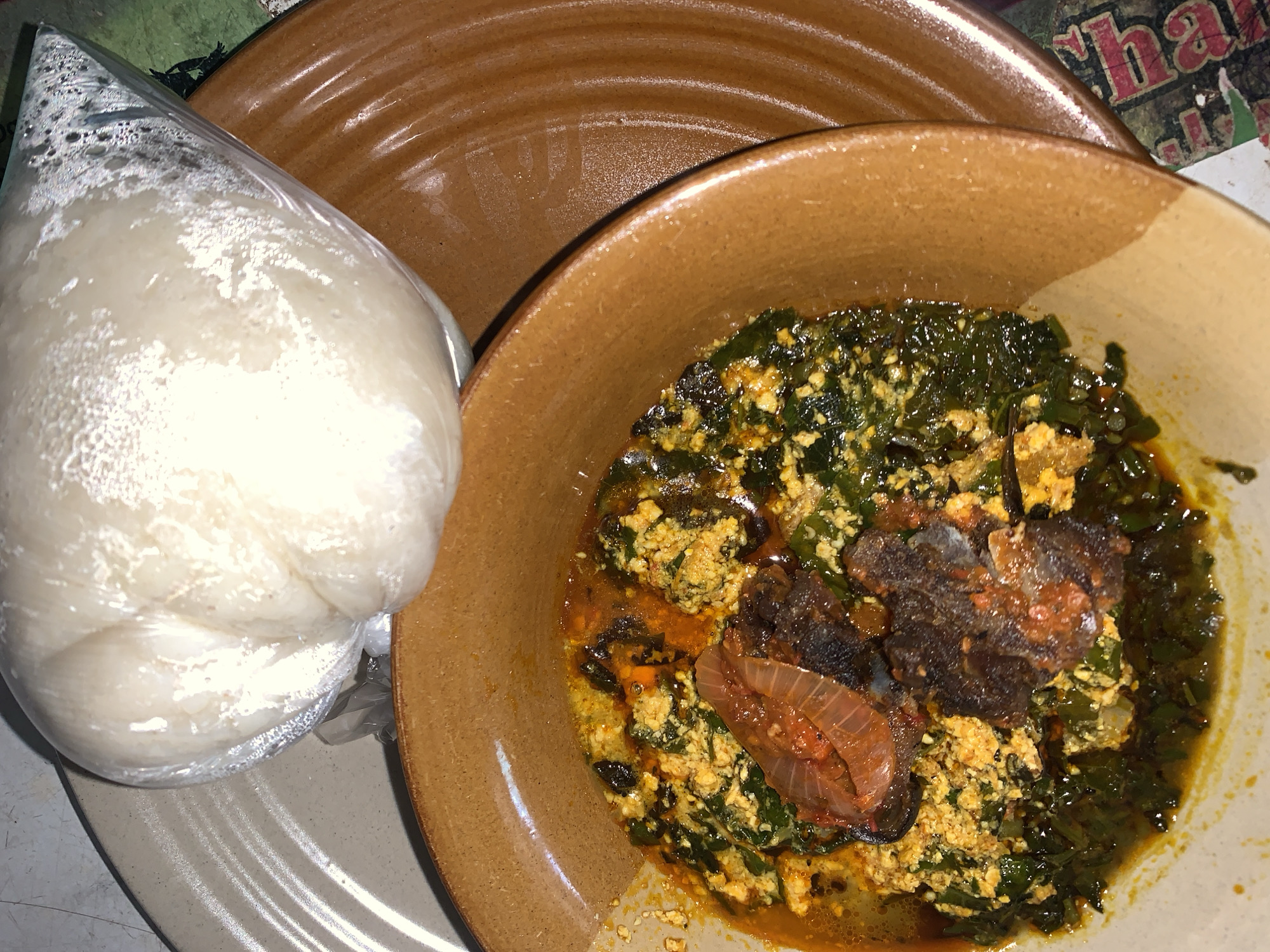 A high-angle close-up shows a meal of eba and soup on a table. Eba, a white, doughy staple food, is wrapped in clear plastic and rests on a plate to the left. Beside it, a bowl of green soup with chunks of fish and onions sits. The scene takes place in New Stadium, Nigeria.