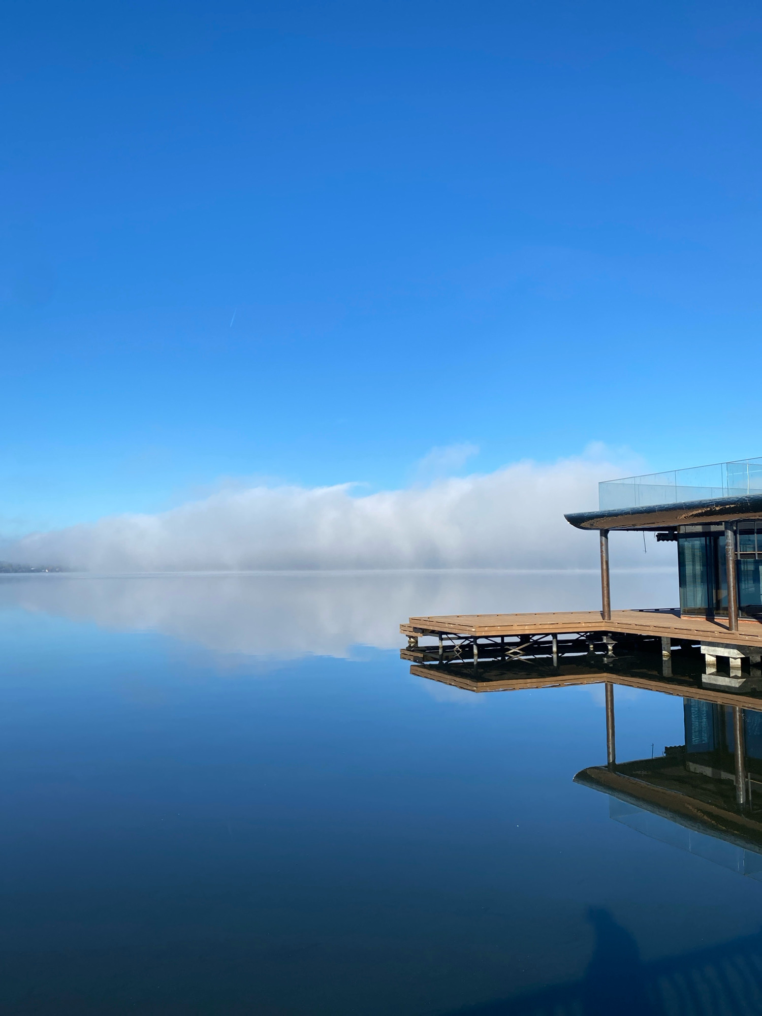 The image depicts a serene lake scene in Votkinsk, Russia. The sky is a clear, bright blue, with a thin line of white clouds stretching across the horizon. The lake's surface is calm and reflective, mirroring the sky and a modern, wooden structure with a glass railing situated on a wooden pier. This structure appears to be a boathouse or a lakeside cafe. The reflection on the water is sharp, capturing the details of the building and its surroundings. A small airplane trail is visible high in the sky. The overall impression is one of tranquility and natural beauty, enhanced by the clean lines of the architecture.