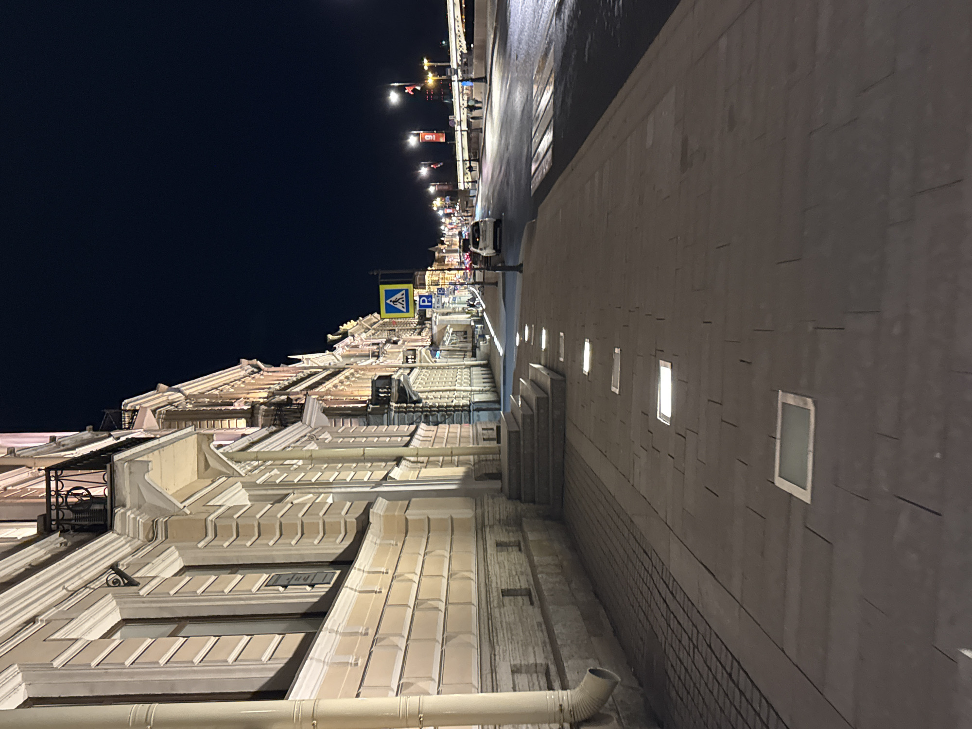 The image shows a nighttime view of a street in Saint Petersburg, Russia. On the left side of the image is the facade of a light-colored building with ornate architectural details, including a rooftop balcony and decorative moldings. A large downspout is visible near the bottom of the frame. The street is seen with several street lights illuminating the area and pedestrian crossing signs are visible. A car is parked along the side of the street. On the right side of the image is a dark gray wall with a pattern of rectangular tiles. Several small, rectangular windows are set into the wall. The dark night sky is visible above the building.