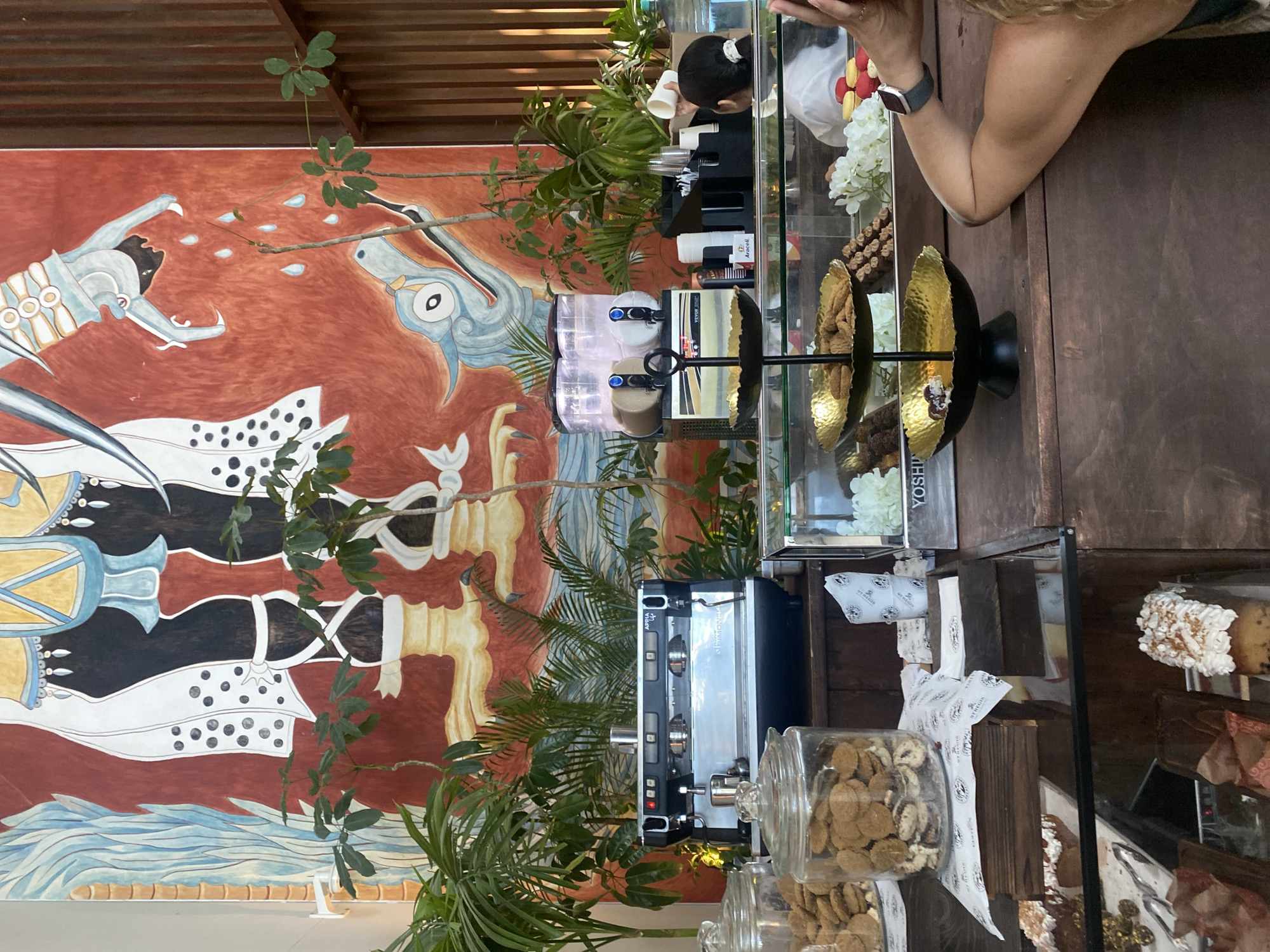 The image shows a detailed view of a commercial interior, likely a cafe or bakery, possibly in Marina Maroma, Mexico. The focus is on the counter area with a variety of displays, equipment, and decorative elements.
**Objects:**
* **Food display:** Glass-fronted display cases with various pastries, cookies in glass jars, and a tiered stand of sweets.
* **Coffee Machine:** A commercial espresso machine is visible.
* **Drink Dispenser:** Beverage dispenser with beverages in clear containers.
* **Mural:** A large, colorful mural depicting stylized figures covers the wall.
* **Plants:** Potted plants are interspersed throughout the counter area, adding a natural element.
* **Serving Items:** Cups, paper towels, dessert holders and other serving items
**People:**
* A person (partial view of arm and watch) is seen near the counter, possibly a customer.
* A person (partial view of hair in a bun) is seen behind the counter, possibly working there.
**Environment:**
* **Location:** The style of mural may suggest Marina Maroma, Mexico.
* **Atmosphere:** The overall atmosphere suggests a modern and stylish cafe or bakery, with an emphasis on visual appeal.
**General Impression:**
A busy and well-decorated commercial space, possibly a cafe or bakery, with a focus on aesthetics and customer experience. The mural, plants, and neatly arranged displays contribute to a visually appealing environment.