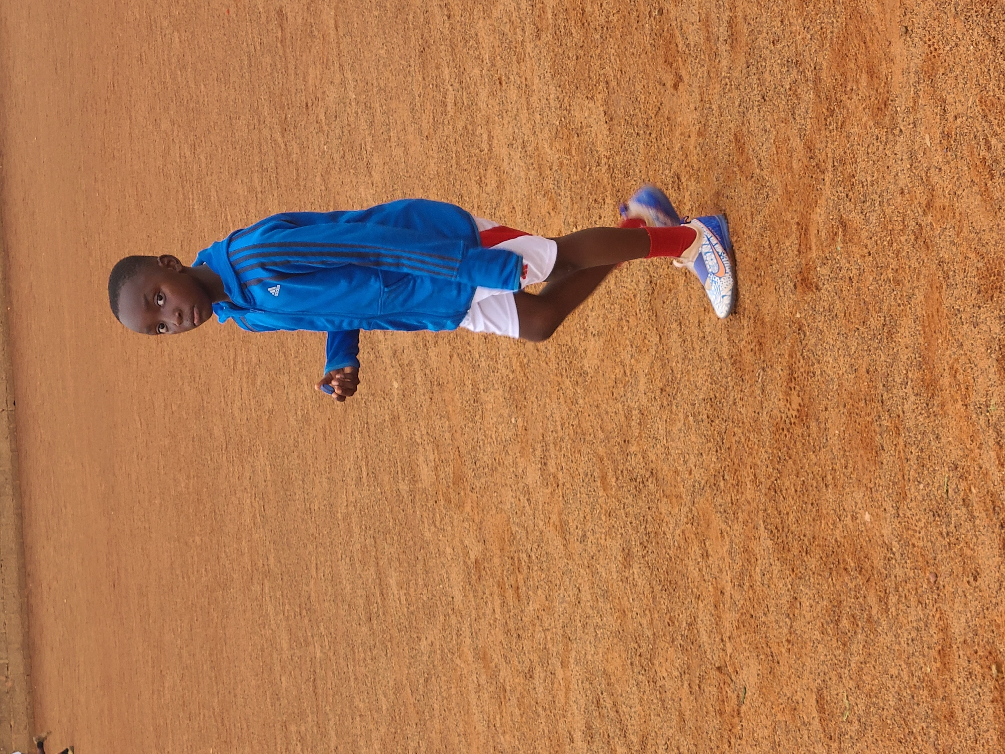 A young person is standing on a reddish-brown surface, likely dirt or clay. The individual is wearing a blue Adidas tracksuit jacket with three stripes on the sleeves, white shorts, red socks, and blue and white shoes. The location appears to be outdoors in Barakin Lerawi, Nigeria.