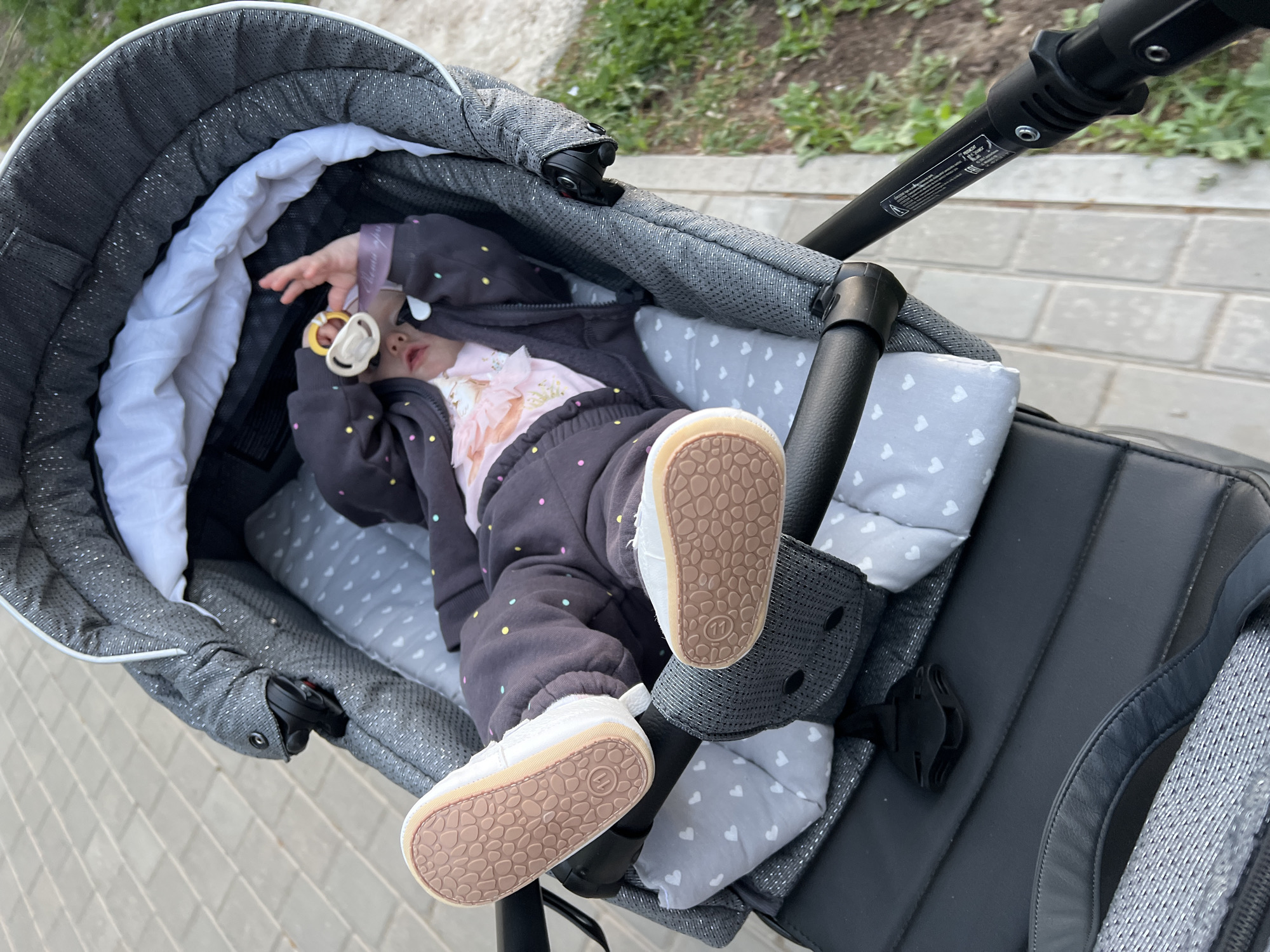 A child lies in a gray baby stroller. The child wears a gray polka-dot sweat suit and white shoes. The soles of the shoes face the camera. A pacifier is in the child's hand. The baby stroller is on a sidewalk next to grass. Location: Chaikovsky, Russia.